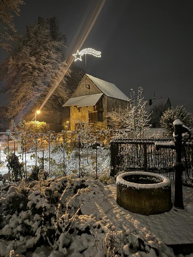 Ein schneebedeckter Garten und ein Brunnen vor einem rustikalen Haus bei Nacht, mit einer leuchtenden Sterndekoration auf dem Dach und warmen Außenlichtern, die die winterliche Szene beleuchten.