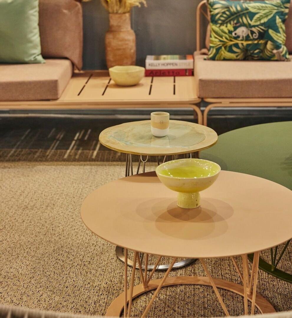 Ein gemütliches Wohnzimmer mit zwei runden Couchtischen mit einer Tasse und einer gelben Schale, einer beigen Bank mit grünen und geblümten Kissen sowie dekorativen Büchern und einer Vase im Hintergrund. Neutrale Töne und warme Beleuchtung schaffen eine entspannte Atmosphäre.