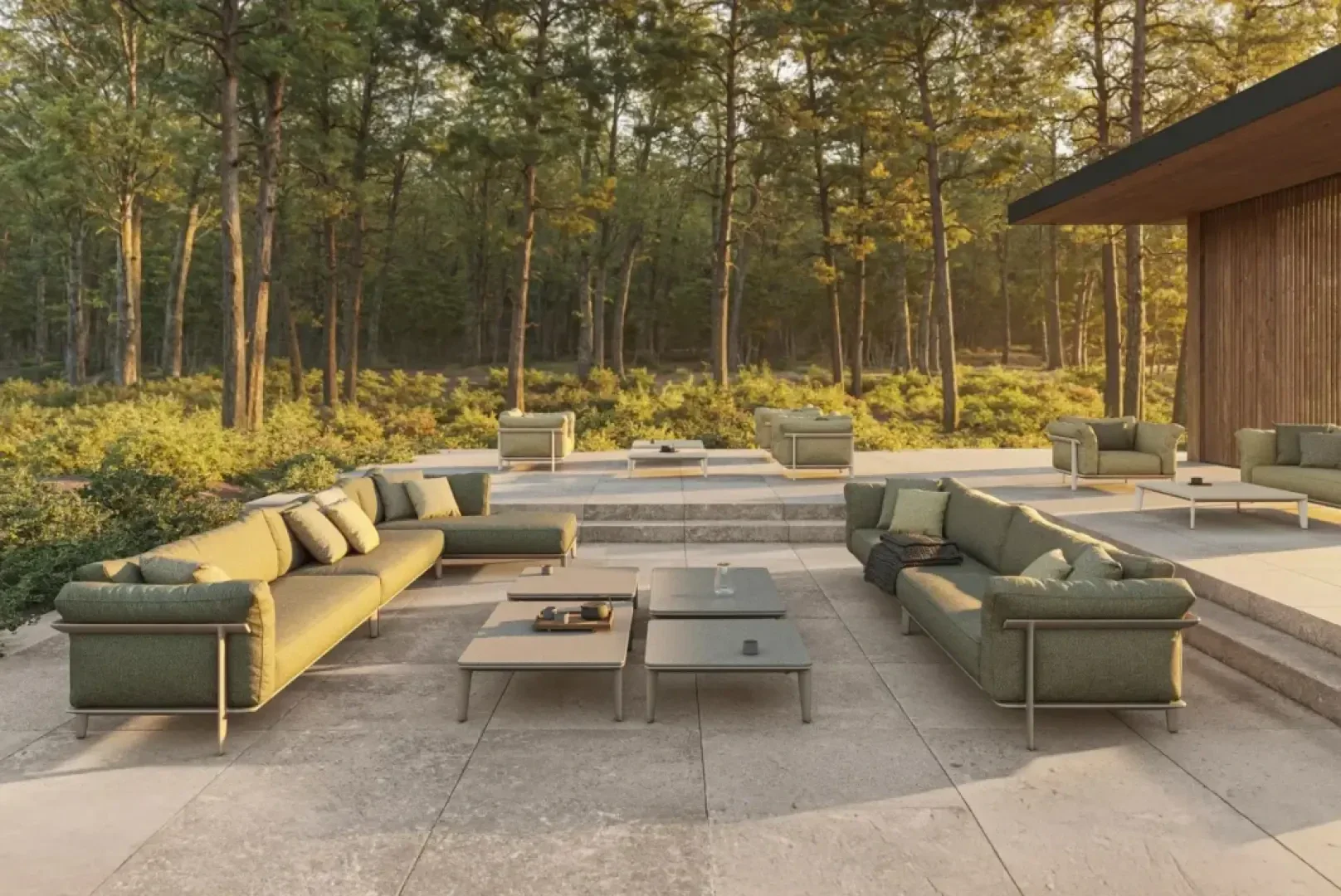 Modern outdoor patio with green sofas, coffee tables, and chairs on stone tiles, surrounded by tall trees and greenery. Sunlight filters through the forest, creating a peaceful, inviting atmosphere.