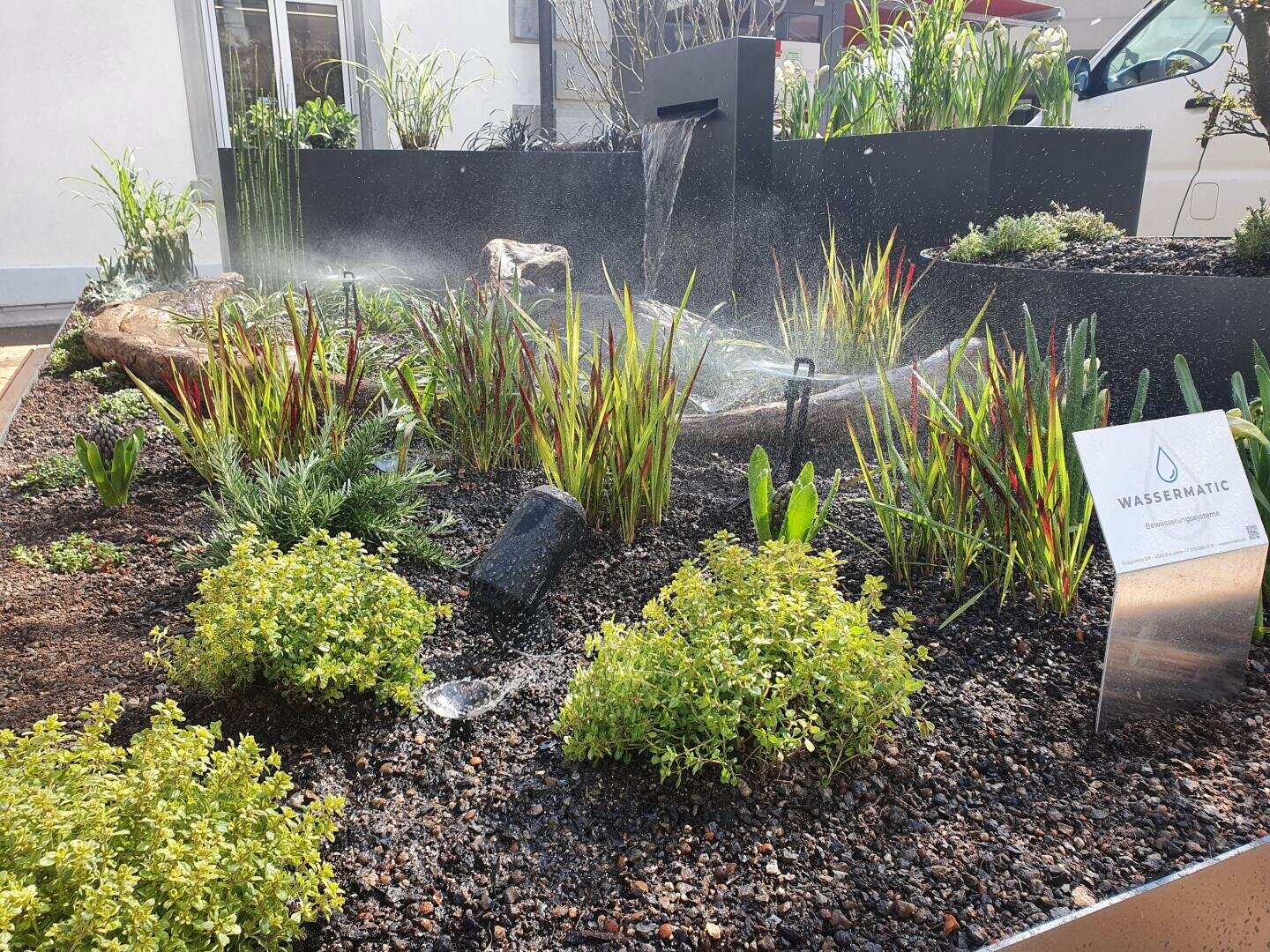 A landscaped garden with green and red ornamental plants is watered with sprinklers. There is a small water feature in the ground in front of the plants and a sign saying WASSERMATIC.