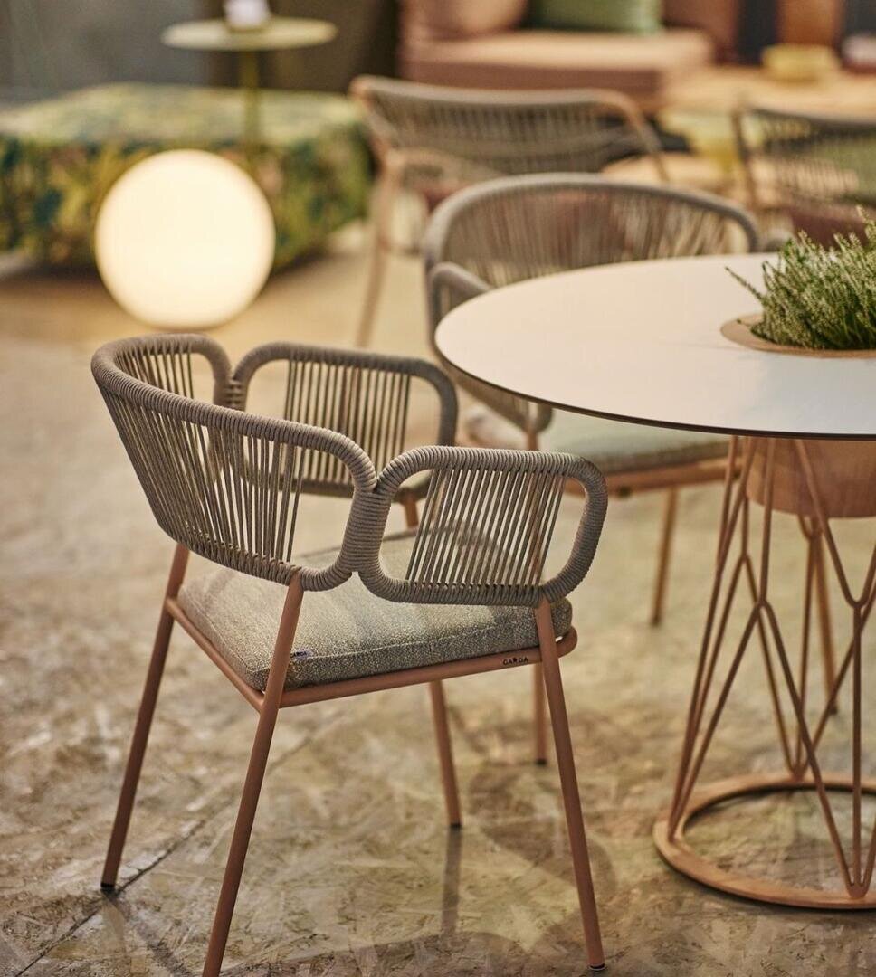 Eine moderne Inneneinrichtung mit einem runden weißen Tisch, umgeben von stilvollen Stühlen mit geflochtenen Rückenlehnen und gepolsterten Sitzen. Im sanft beleuchteten Hintergrund sind eine leuchtende Kugel und eine gemusterte Ottomane zu sehen.