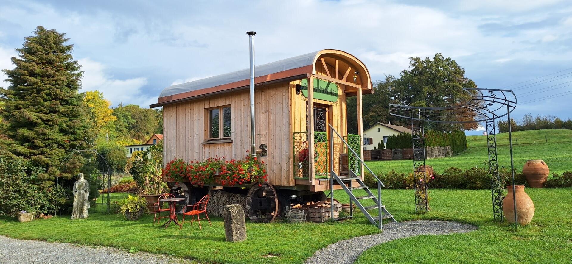 Ein kleines Holzhäuschen auf Rädern mit einem geschwungenen Dach und einem Metallschornstein steht in einem grünen Garten. Rote Blumen säumen die Veranda, und eine Metallleiter führt zur Tür hinauf. Stühle im Freien und Topfpflanzen umgeben das Haus.