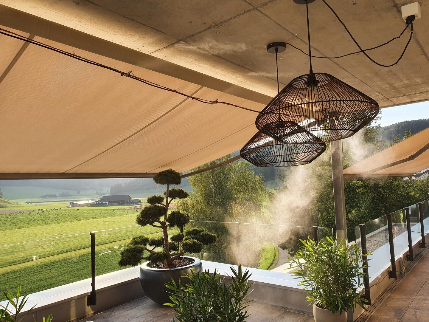 A modern terrace with hanging lamps, potted plants and awnings offers a view of green fields and trees. Sunlight filters in while mist rises from a luminaire, creating a tranquil outdoor atmosphere.