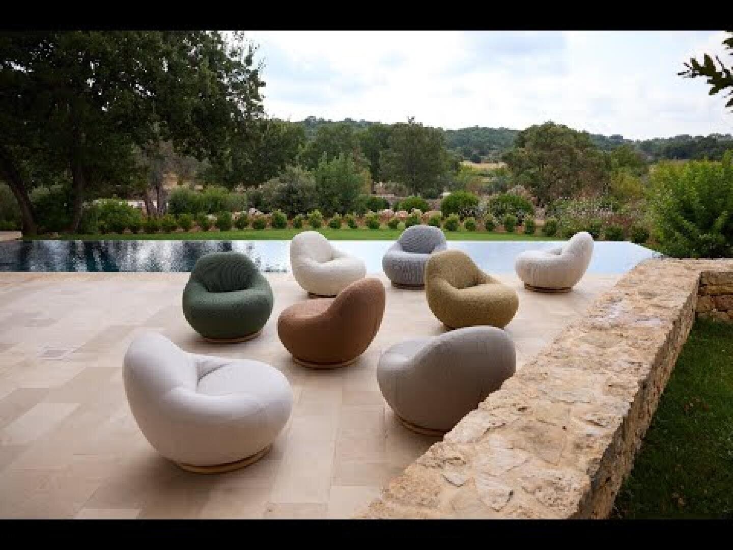 A terrace with round, cushioned sun loungers in different colors stands against a stone wall, overlooking a pool and lush greenery with trees and hills in the background.