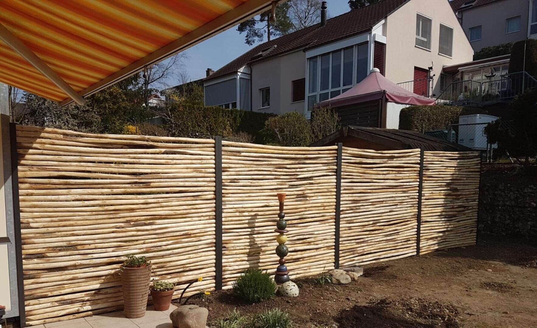 Ein Hinterhof mit einem hohen Sichtschutzzaun aus horizontalen Holzstämmen, Gartentöpfen, einer bunten, gestapelten Skulptur und Häusern mit Schrägdächern im Hintergrund.