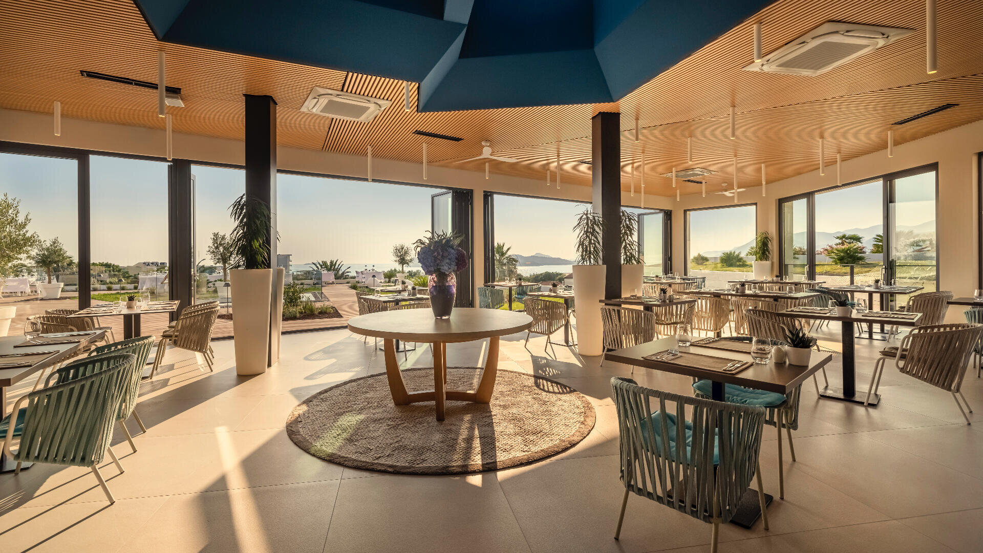 Ein modernes, lichtdurchflutetes Restaurant mit großen Fenstern, eleganten Tischen und Stühlen, einem runden Tisch mit einem Blumenarrangement in der Mitte und einem malerischen Blick ins Grüne und auf das Wasser in der Ferne.