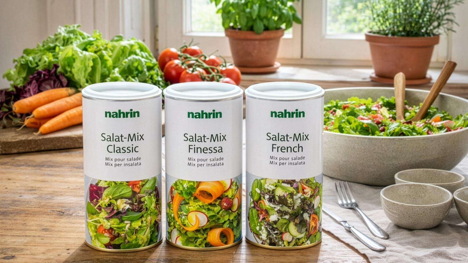 Three cans of Nahrin salad mix (Classic, Finessa, French) on a wooden table, surrounded by fresh vegetables, a salad bowl, plates and cutlery, with a bright window and potted plants in the background.