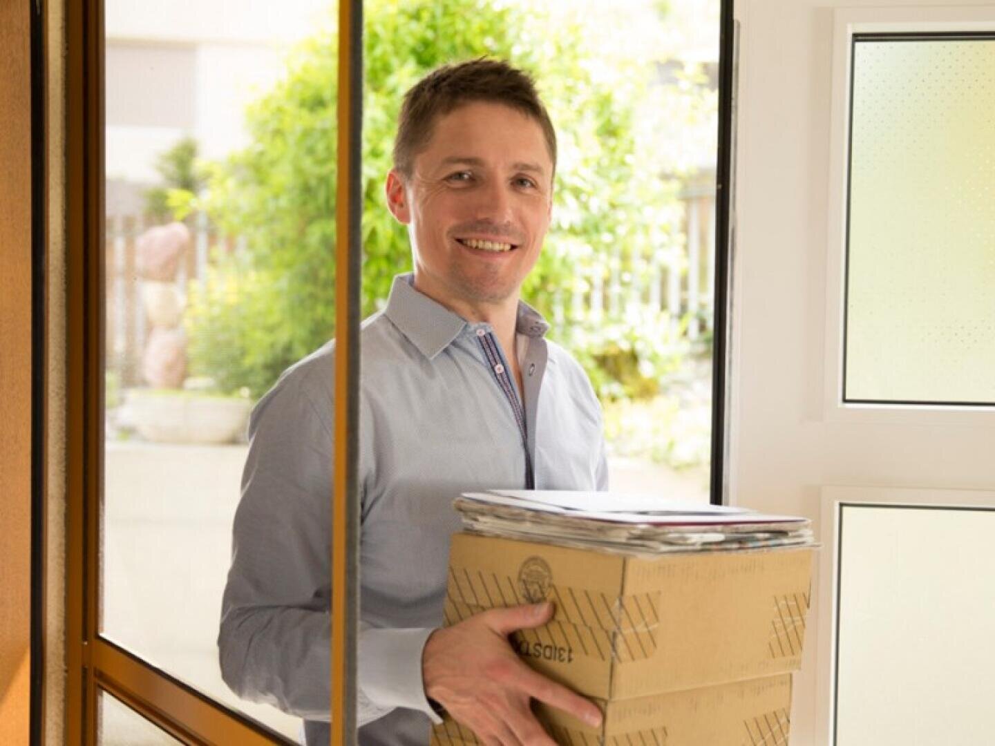 Ein lächelnder Mann in einem hellblauen Hemd steht an einer Tür und hält zwei Pappkartons und etwas Post in der Hand. Hinter ihm sind helles Tageslicht und Grünflächen zu sehen.