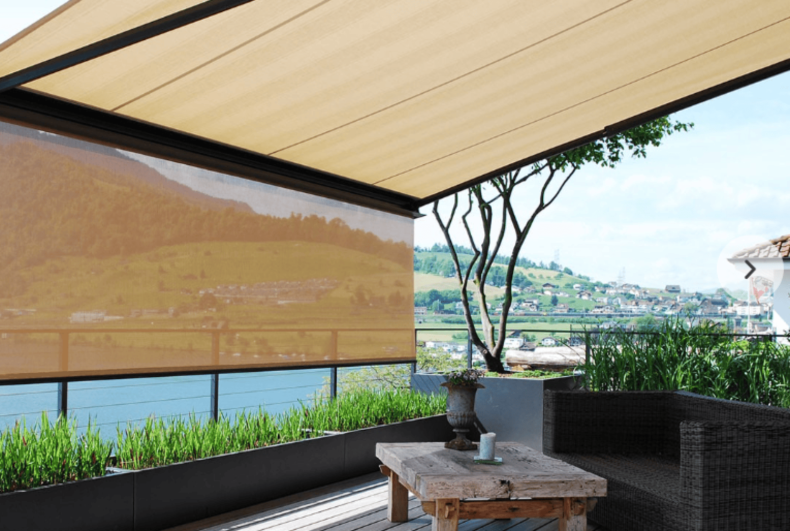 Eine schattige Außenterrasse mit Holztisch, Korbsofa, Topfpflanzen und Blick auf einen Fluss, grüne Hügel und ein Dorf im Hintergrund unter einer braunen, ausfahrbaren Markise.