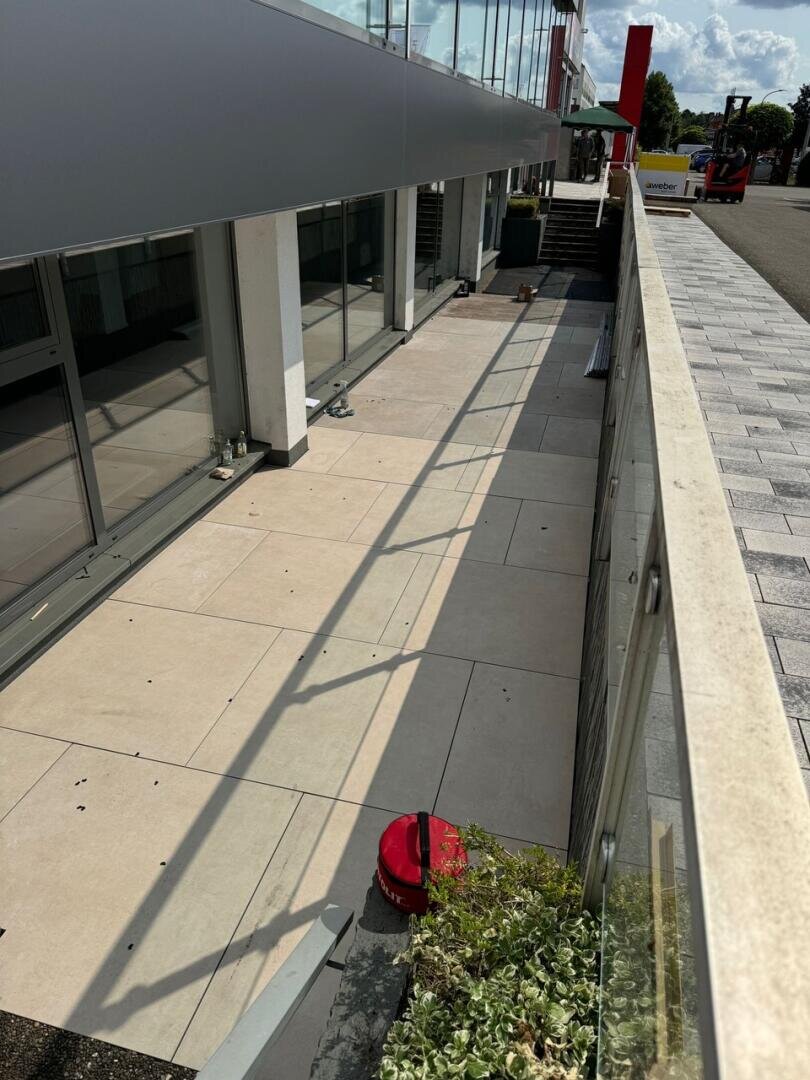 Ein Glasgeländer blickt auf eine große, leere geflieste Terrasse neben einem modernen Gebäude mit großen Fenstern. Ein roter Eimer steht am Rande der Terrasse, und im Vordergrund sind einige Topfpflanzen zu sehen.