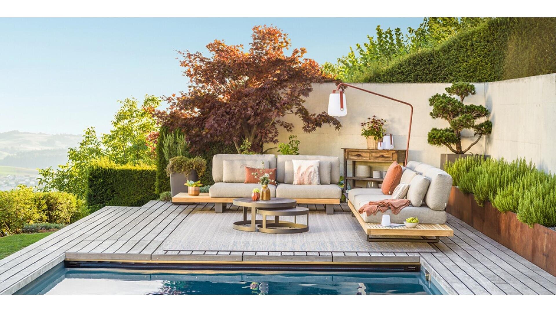 Modern outdoor patio with gray cushioned sofas, round coffee tables, plants, and decor set on a wooden deck beside a swimming pool, surrounded by greenery and landscaped garden.