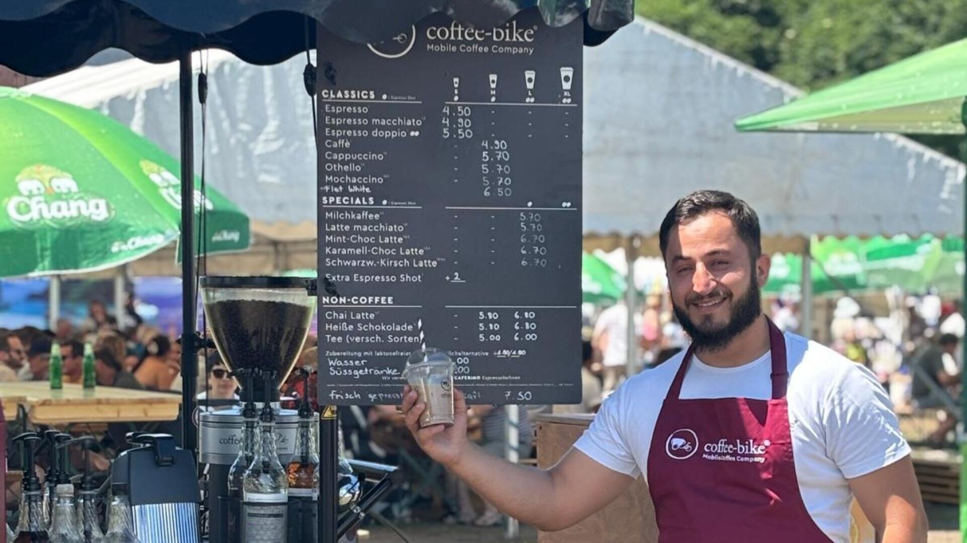 Ein lächelnder Barista in einer kastanienbraunen Schürze steht an einem Kaffeestand und hält ein Getränk in der Hand. Hinter ihm zeigt eine große schwarze Tafel die verschiedenen Getränke an, während im Hintergrund ein belebter Markt zu sehen ist.