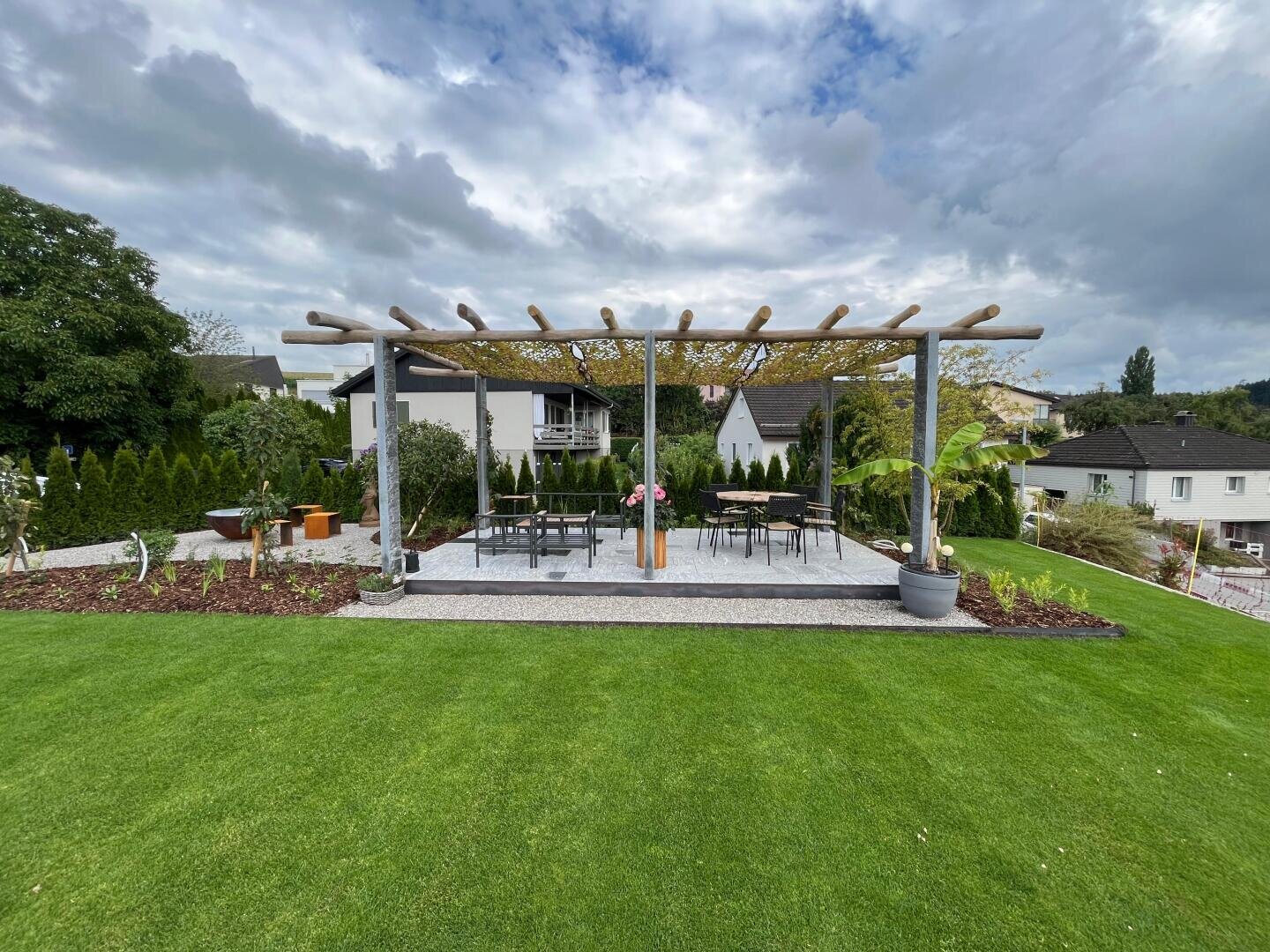 Eine hölzerne Pergola mit einem teilweise überdachten Dach steht auf einer Terrasse, die von Grünflächen umgeben ist. Darunter stehen Tische und Stühle, die mit Topfpflanzen und einer Blumenvase geschmückt sind. Im Hintergrund sind Häuser und Bäume zu sehen.