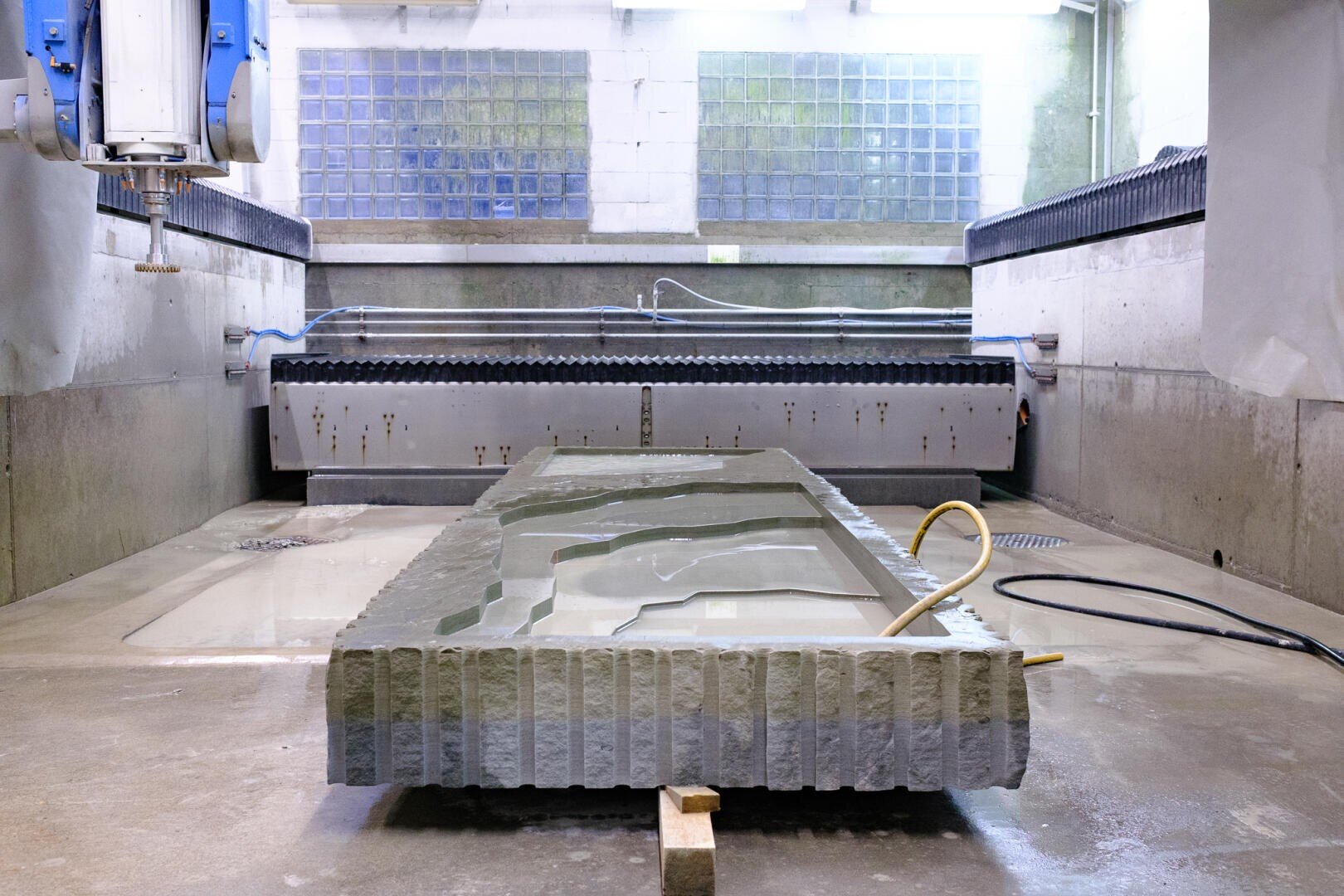 Eine große Steinplatte mit verschlungenen Rillen liegt in einer Industriehalle, umgeben von Betonwänden und Glasblöcken. Rohre, Schläuche und Maschinen sind zu sehen, was darauf schließen lässt, dass die Steinbearbeitung oder die Herstellung im Gange ist.