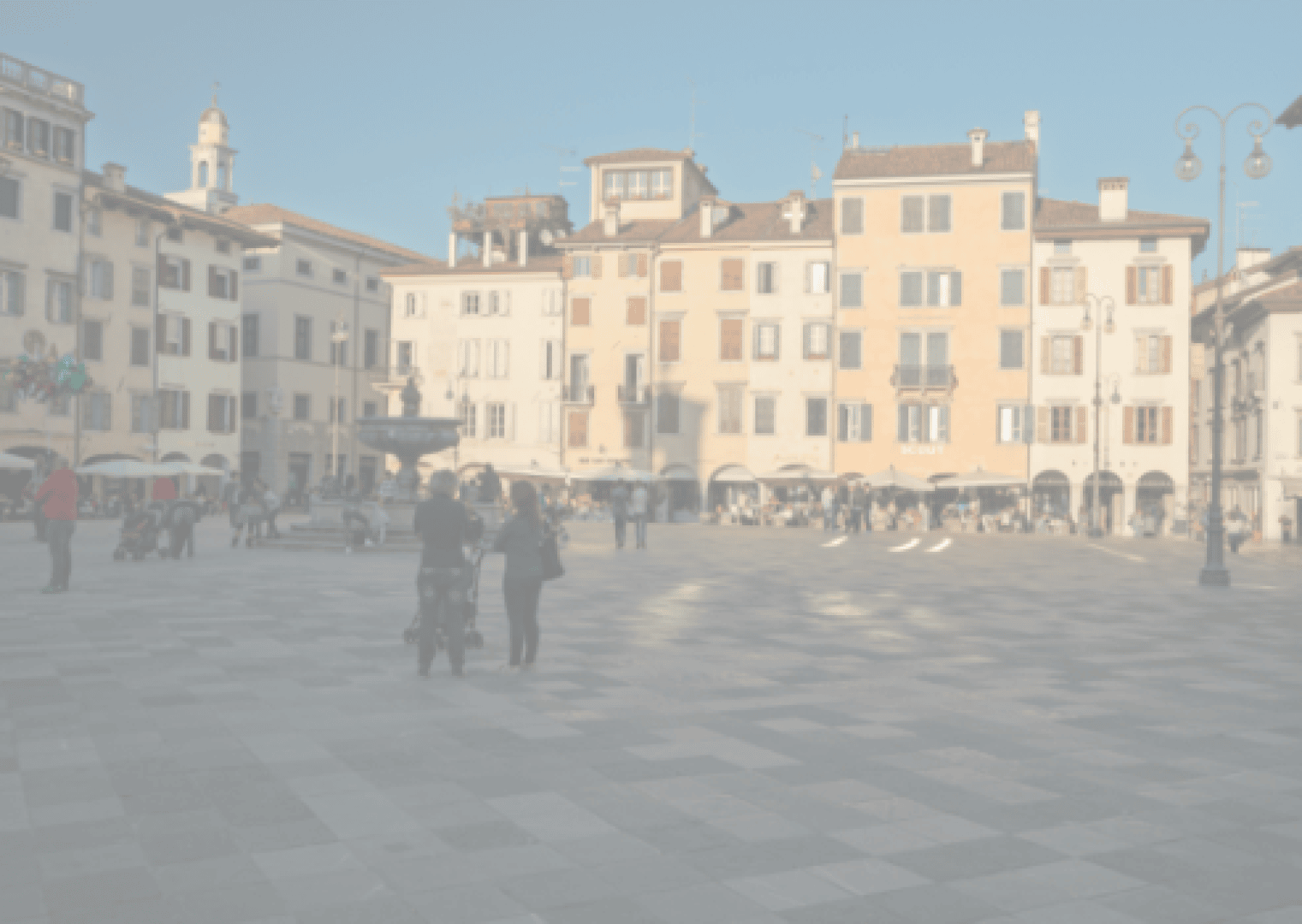 Die Menschen gehen spazieren und versammeln sich auf einem geräumigen, sonnenbeschienenen Stadtplatz mit gemusterten Pflastersteinen, umgeben von farbenfrohen historischen Gebäuden und einem zentralen Springbrunnen. Einige Menschen sitzen auf Bänken oder schlendern über die Freifläche.