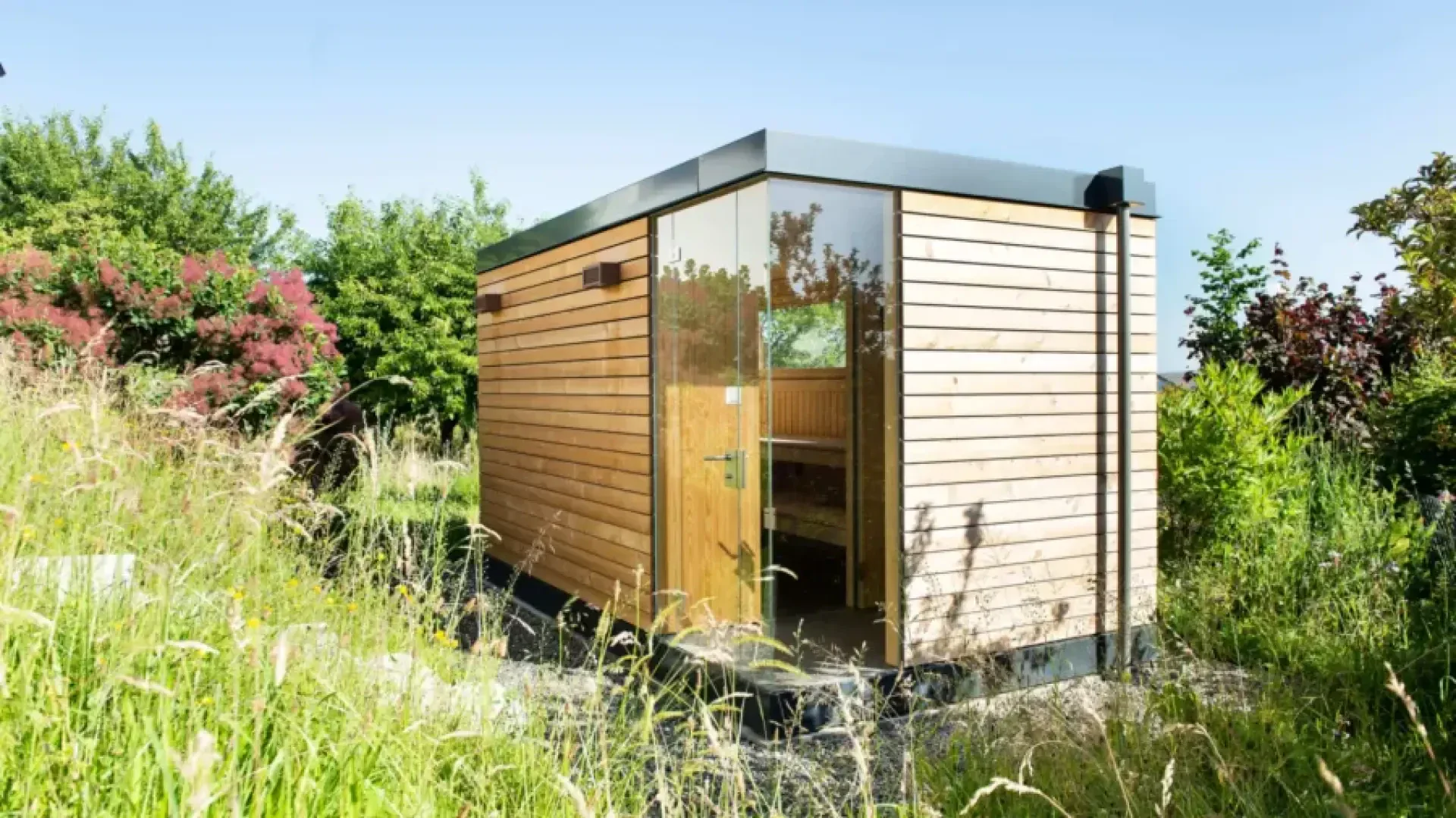 Eine moderne, kleine Holzhütte mit vertikaler Verkleidung steht inmitten von hohem Gras und üppigem Grün unter einem klaren blauen Himmel. Die Hütte hat große Glastüren und minimalistische Designmerkmale.