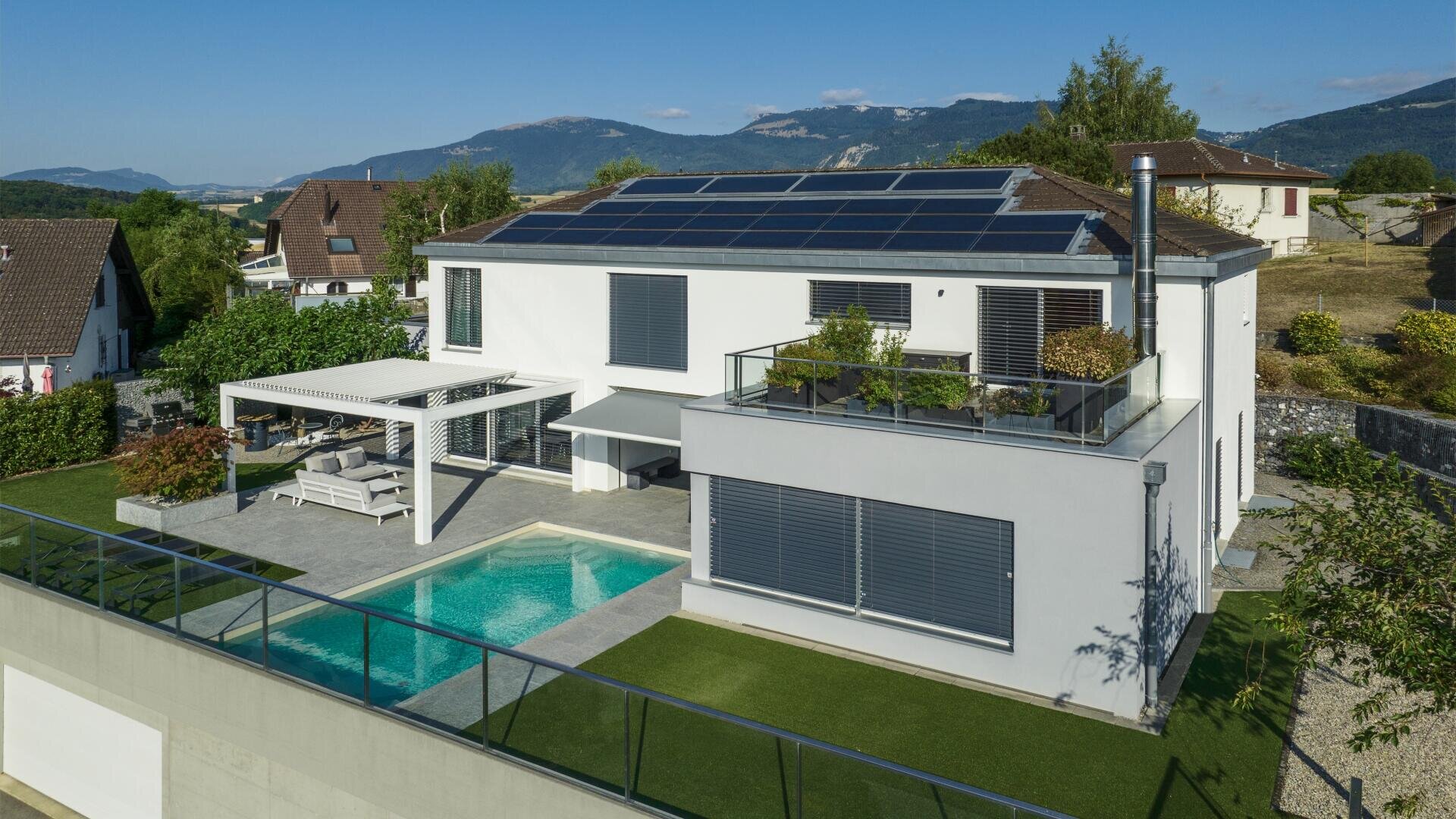 Modern white two-story house with large solar panels on the roof, a swimming pool, outdoor seating area under a pergola, and a balcony with plants, set in a residential neighborhood with mountains in the background.