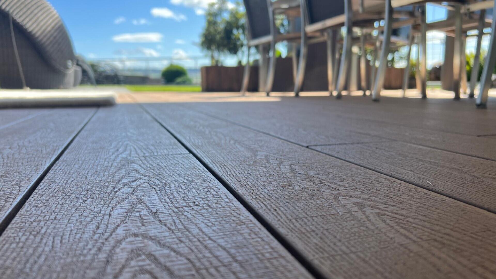 Close-up view of textured wooden deck flooring with outdoor dining chairs and table in the background, blurred trees, and a bright blue sky with clouds.