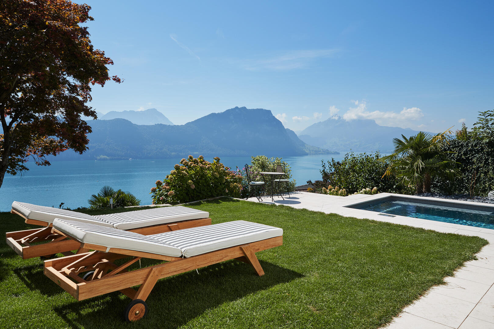 Zwei hölzerne Liegestühle mit weißen Kissen stehen auf einer grasbewachsenen Terrasse mit Blick auf einen See und die Berge. Ein kleiner Pool und ein Tisch mit Stühlen sind in der Nähe, umgeben von Bäumen und Pflanzen unter einem klaren blauen Himmel.