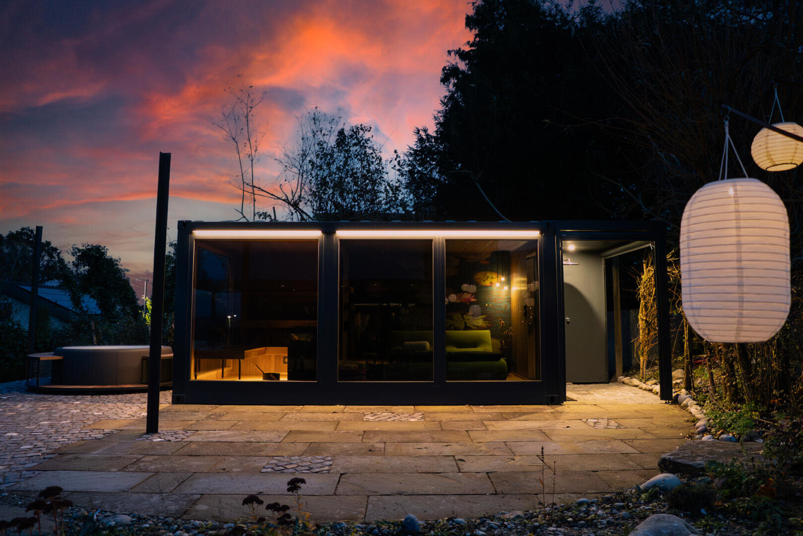 A modern, glass hut is illuminated at dusk, with warm light inside and a glowing sunset sky in the background. Paper lanterns hang to the right, and a stone terrace leads to the entrance. The scene is surrounded by trees.