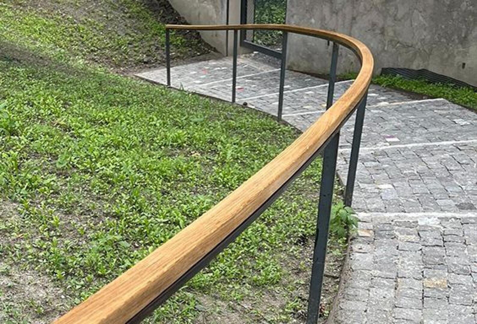 A gently curving wooden handrail follows a path paved with stones, bordered on one side by green grass and on the other by a concrete wall. The path leads to a glass door in the background.