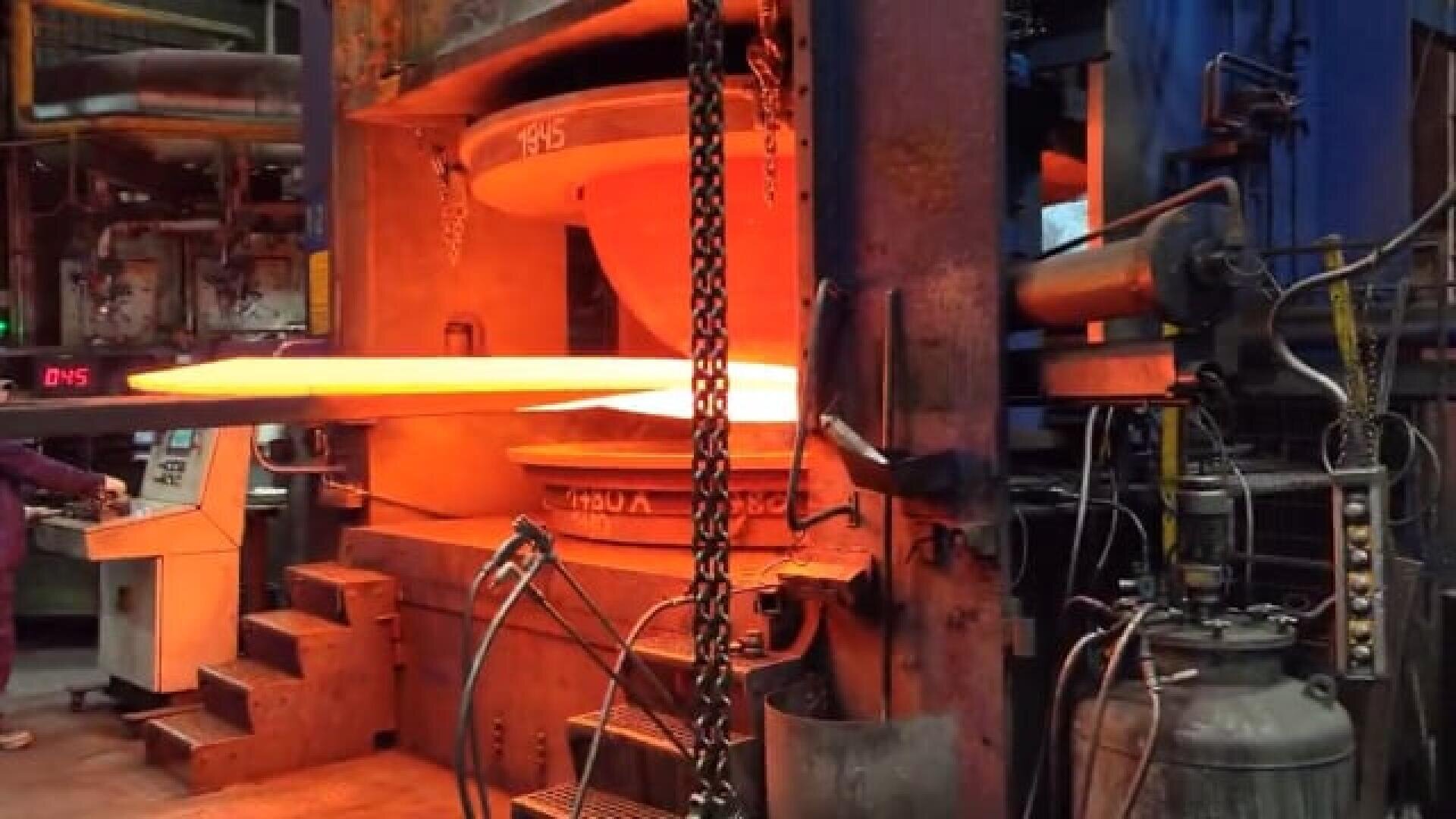 Ein glühendes, heißes Metallwerkstück wird in einer großen Industriepresse in einer Fabrik geschmiedet, umgeben von Maschinen, Schalttafeln und Metallketten.