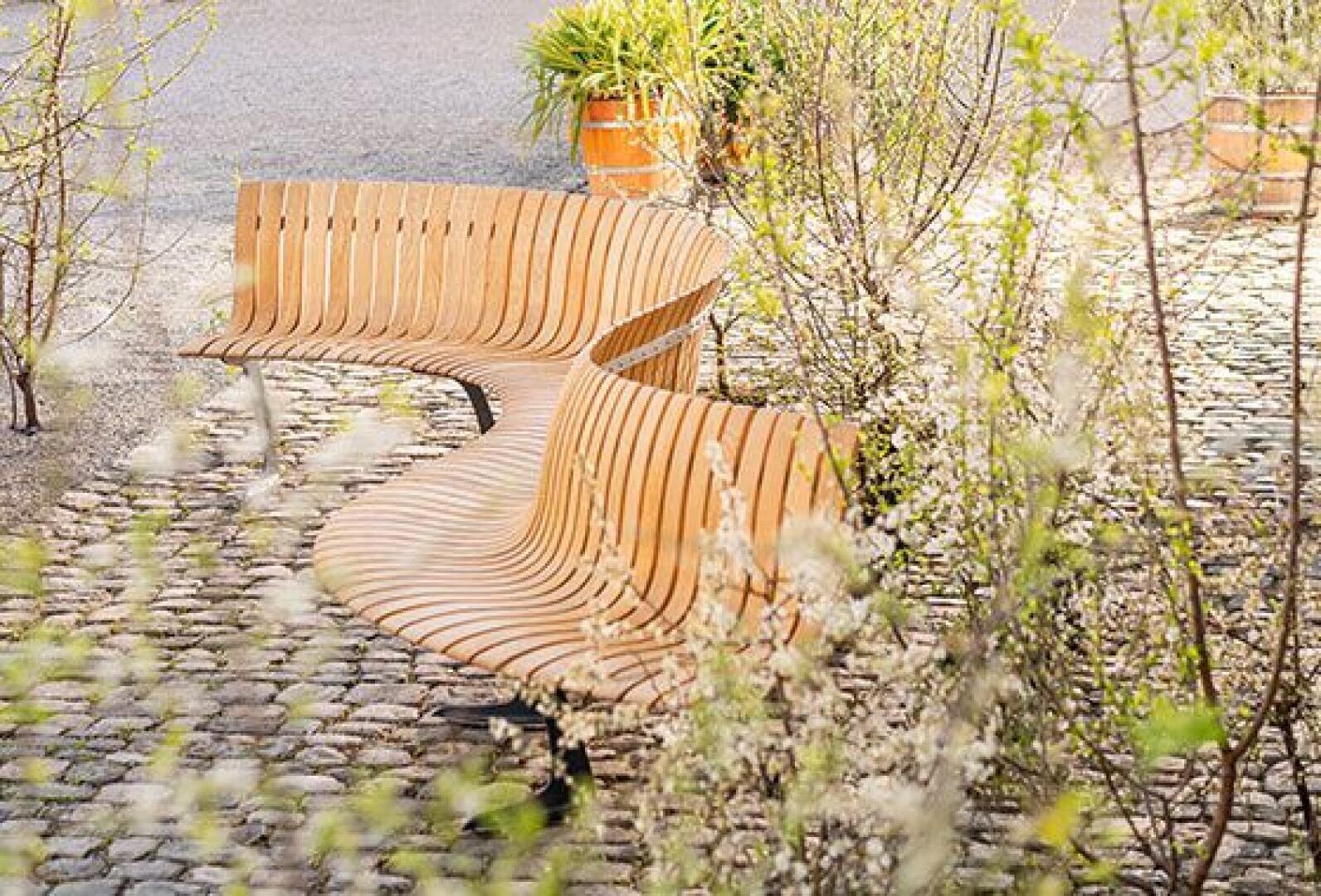 Geschwungene Holzbänke auf einem Kopfsteinpflasterweg, umgeben von blühenden Sträuchern und einer Topfpflanze im Hintergrund. Die Szene wirkt hell und einladend und suggeriert eine friedliche Umgebung im Freien.