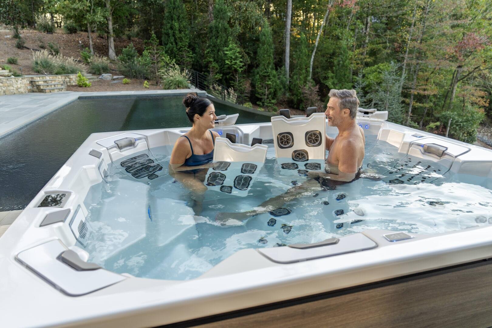 Ein Mann und eine Frau entspannen und unterhalten sich in einem modernen Whirlpool im Freien in der Nähe eines Pools, umgeben von Bäumen und Grünpflanzen im Hintergrund.