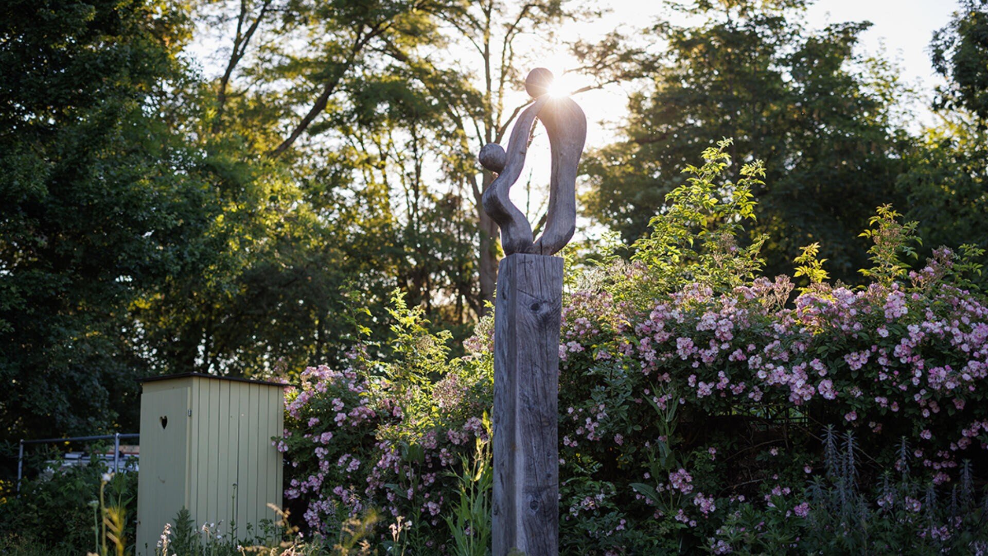 Eine Holzskulptur mit zwei abstrakten menschlichen Figuren steht in einem Garten mit rosa Blumen, hohem Grün und Sonnenlicht, das durch die Bäume im Hintergrund fällt.