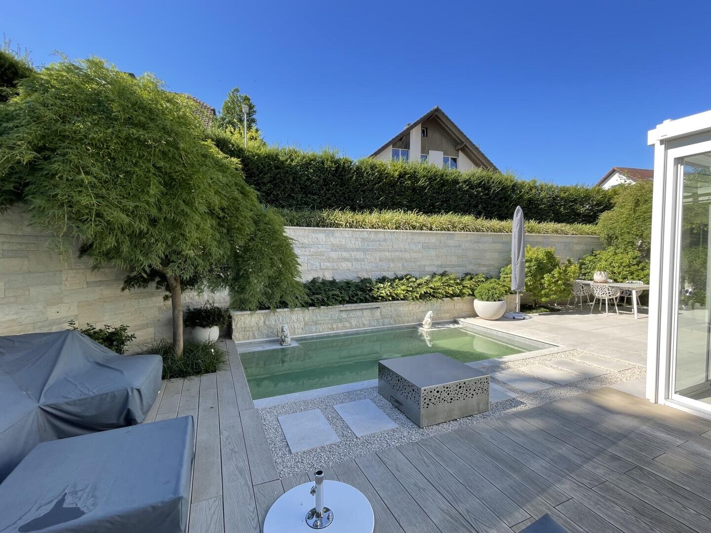 Moderner Hinterhof mit einem kleinen rechteckigen Pool, umgeben von Stein und Grün. Es gibt Sitzgelegenheiten im Freien, einen Tisch mit Stühlen und einen geschlossenen Sonnenschirm unter einem klaren blauen Himmel.