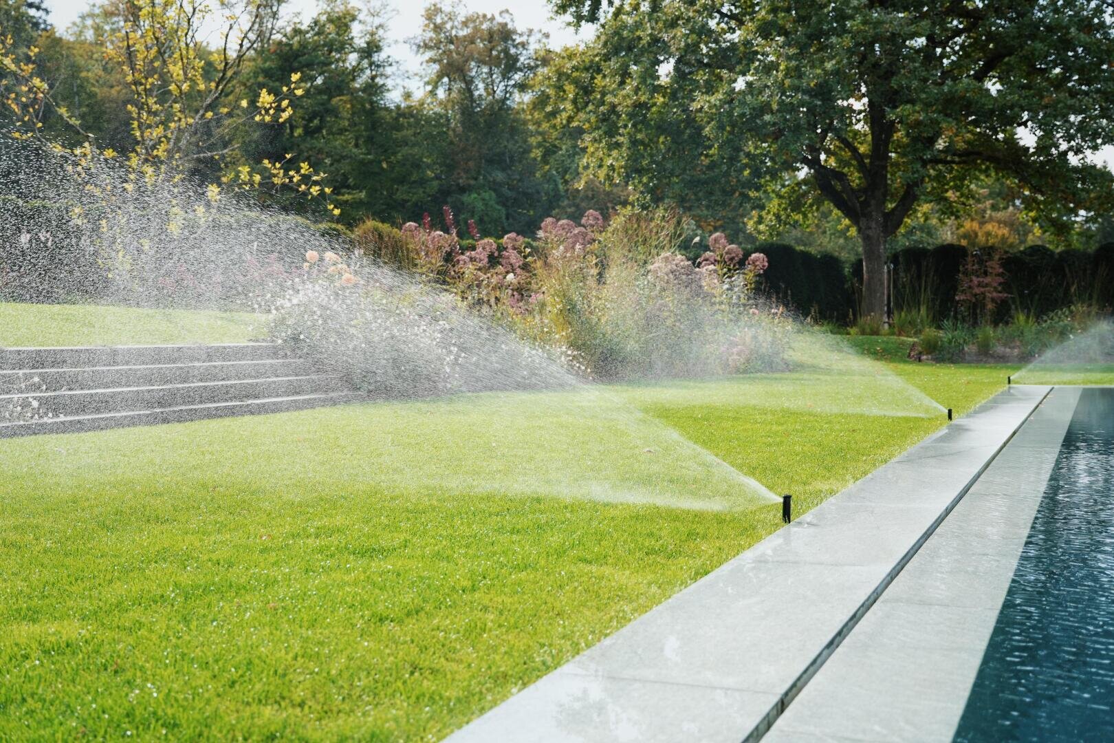 Sprinkleranlagen bewässern einen üppigen grünen Rasen neben einem Pool, mit Gartentreppen und blühenden Pflanzen im Hintergrund und schattenspendenden Bäumen.