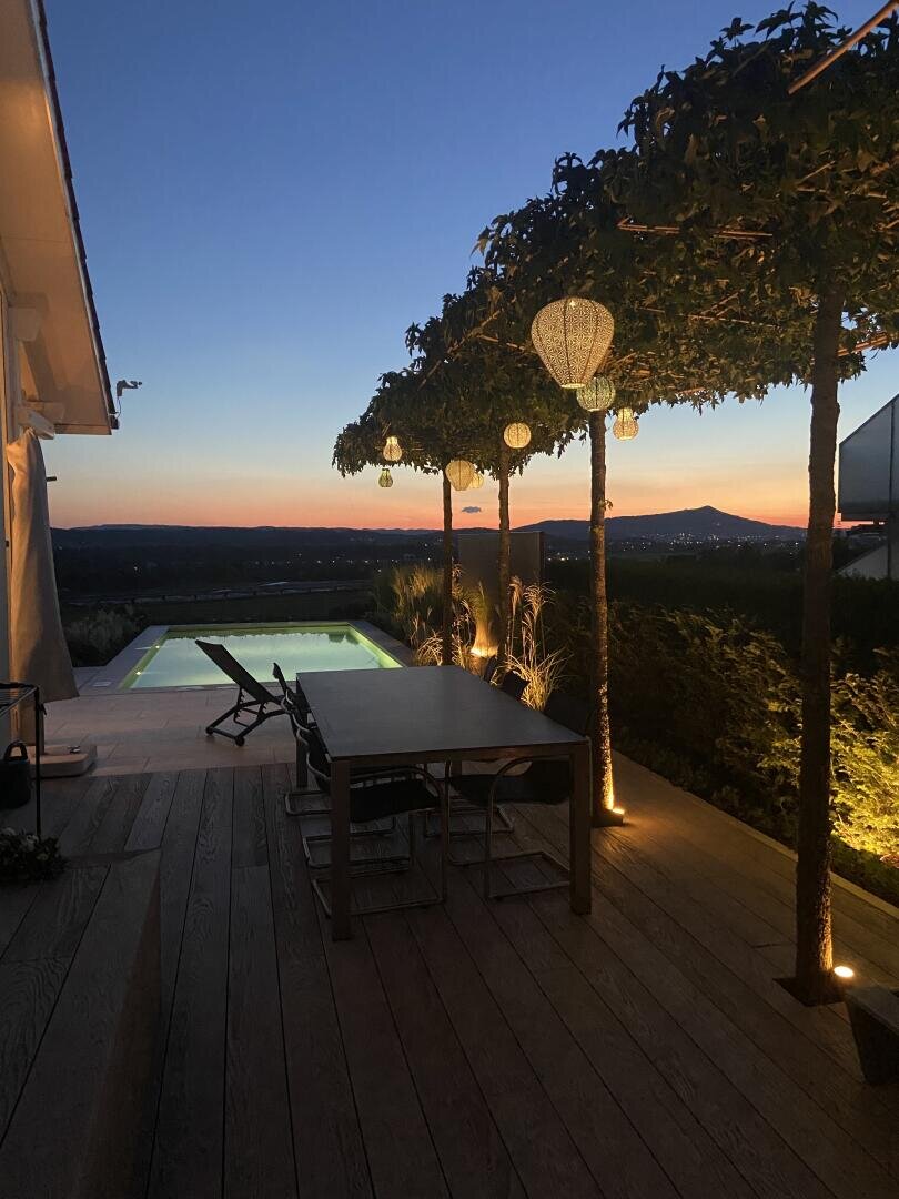 Eine moderne Außenterrasse bei Sonnenuntergang mit Esstisch, Stühlen, Lichterketten, die von Pergolen herabhängen, und einem leuchtenden Pool mit Blick auf eine malerische Landschaft mit Bergen und einem klaren Himmel.