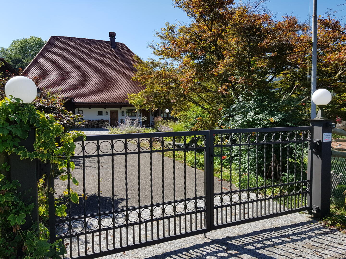Ein schwarzes Metalltor mit kreisförmigen Mustern steht vor einem Haus mit steilem, rotem Ziegeldach, umgeben von grünen Bäumen und Blättern unter einem klaren blauen Himmel.