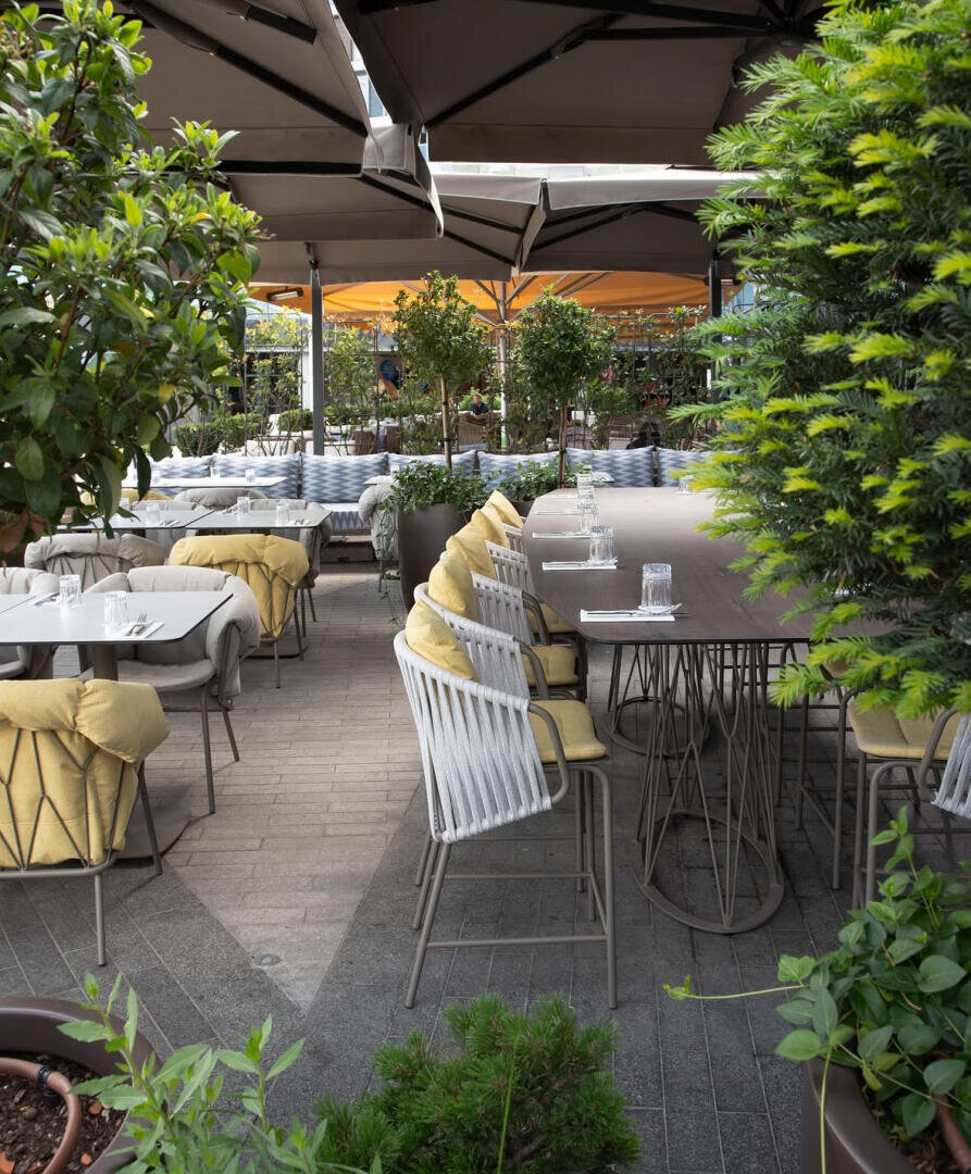 Terrasse des Außenrestaurants mit ordentlich aufgestellten Tischen und Stühlen, gepolsterten Sitzen mit gelben Kissen, umgeben von Grünpflanzen und beschattet von großen Sonnenschirmen. Es sind keine Menschen anwesend.
