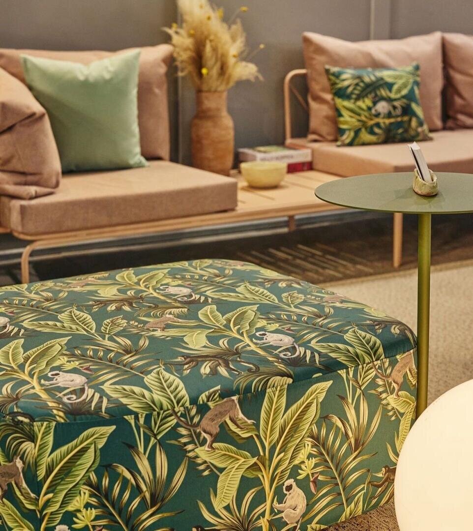 Ein gemütliches Wohnzimmer mit neutralen Sofas, grünen und beigen Kissen, einem dekorativen Pflanzenarrangement und einer Ottomane mit einem tropischen Blatt- und Affenmuster neben einem runden grünen Beistelltisch, auf dem ein Telefon liegt.