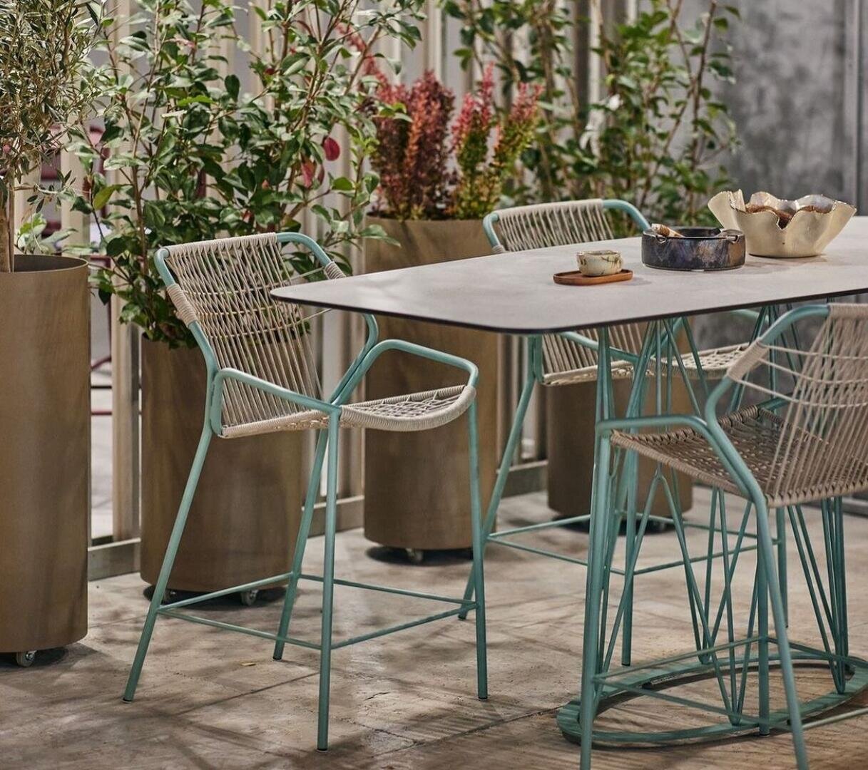 Eine moderne Außenterrasse mit einem hellen Tisch, passenden geflochtenen Stühlen mit Metallbeinen und hohen Topfpflanzen im Hintergrund, die eine gemütliche und stilvolle Gartenatmosphäre schaffen.