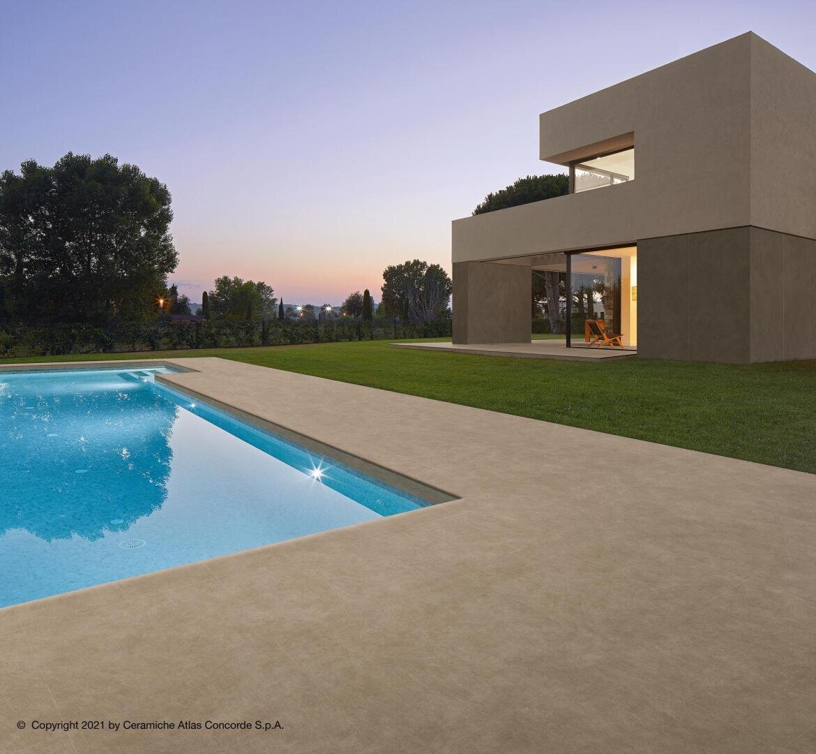 Ein modernes, minimalistisches Haus mit großen Fenstern blickt bei Sonnenuntergang auf einen klaren blauen Swimmingpool, umgeben von einem gepflegten Rasen und hohen Bäumen im Hintergrund.