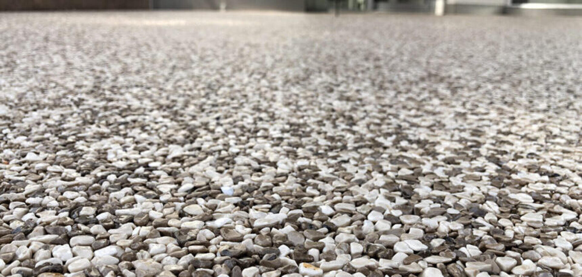 Close-up view of a pebble stone floor with small, smooth, white and brown stones, creating a textured surface. The background appears blurred, showing an indoor or outdoor space.