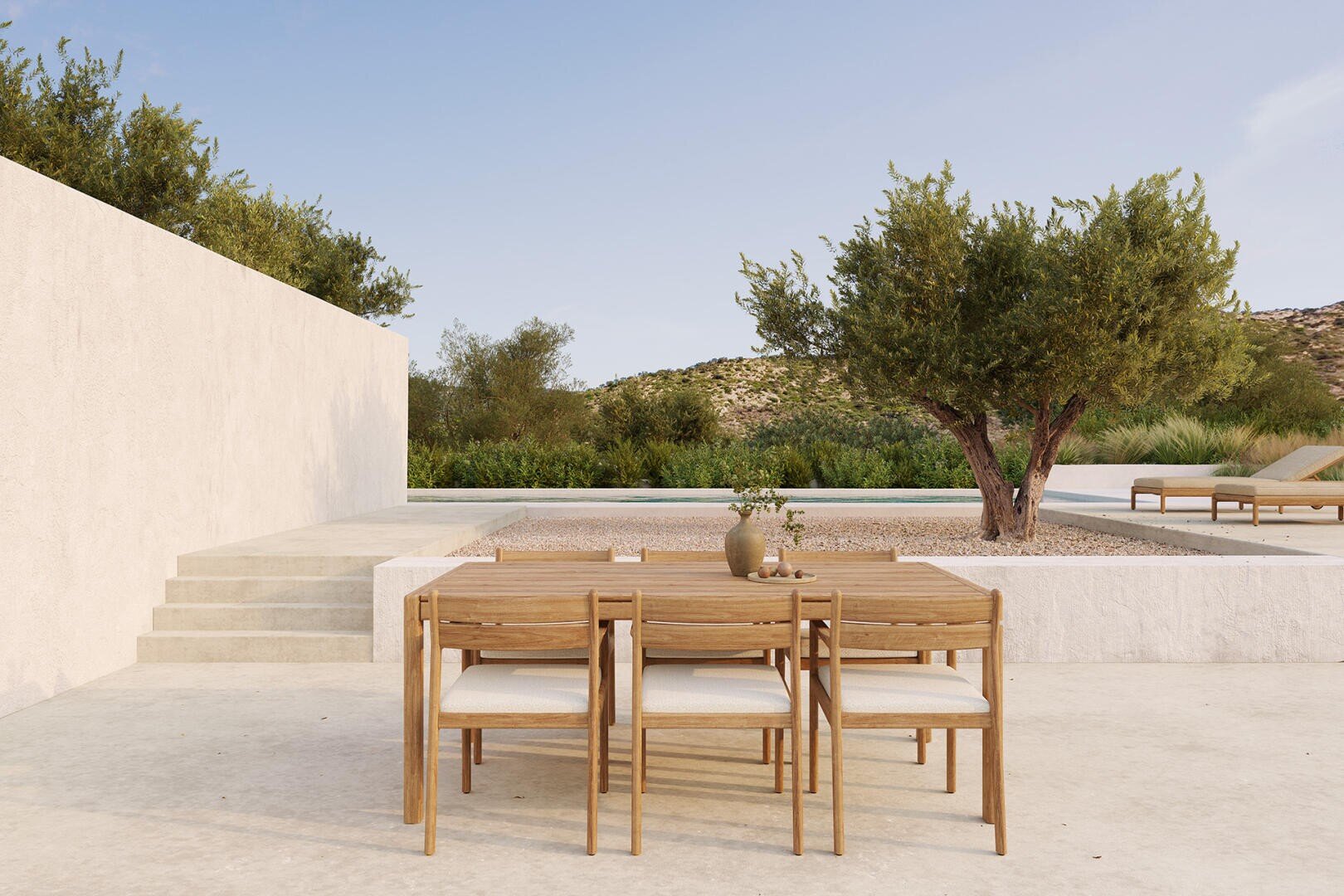 Minimalistische Außenterrasse mit Esstisch und Stühlen aus Holz auf einer hellen Steinfläche, in der Nähe eines kleinen Baums, einer Treppe und eines Pools mit Liegestühlen, umgeben von viel Grün unter einem klaren Himmel.