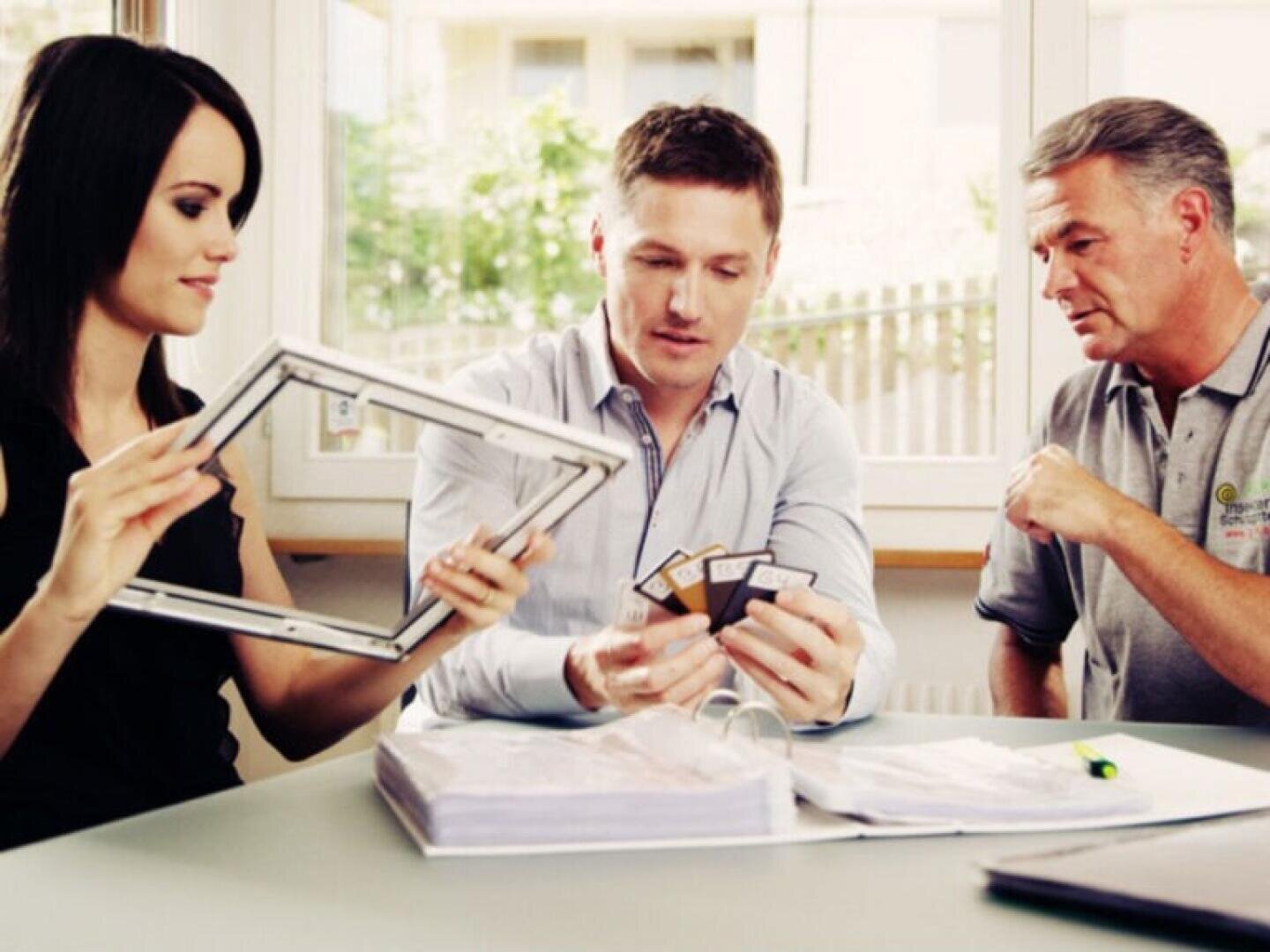Drei Personen sitzen an einem Tisch und diskutieren über die Möglichkeiten von Fensterrahmen. Die Frau hält einen Musterrahmen in der Hand, während die beiden Männer Farbmuster prüfen. Auf dem Tisch vor ihnen sind Papiere und Unterlagen ausgebreitet.