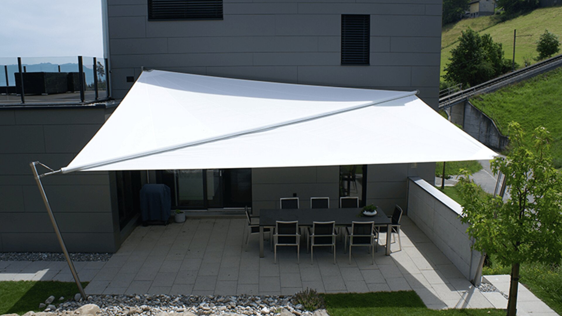 Eine moderne Terrasse mit einem rechteckigen Tisch und acht Stühlen wird von einem großen, weißen, dreieckigen Sonnensegel beschattet, das an einem grauen Gebäude angebracht ist. Die Terrasse ist von kleinen Steinen, Gras und einem nahen Baum umgeben.