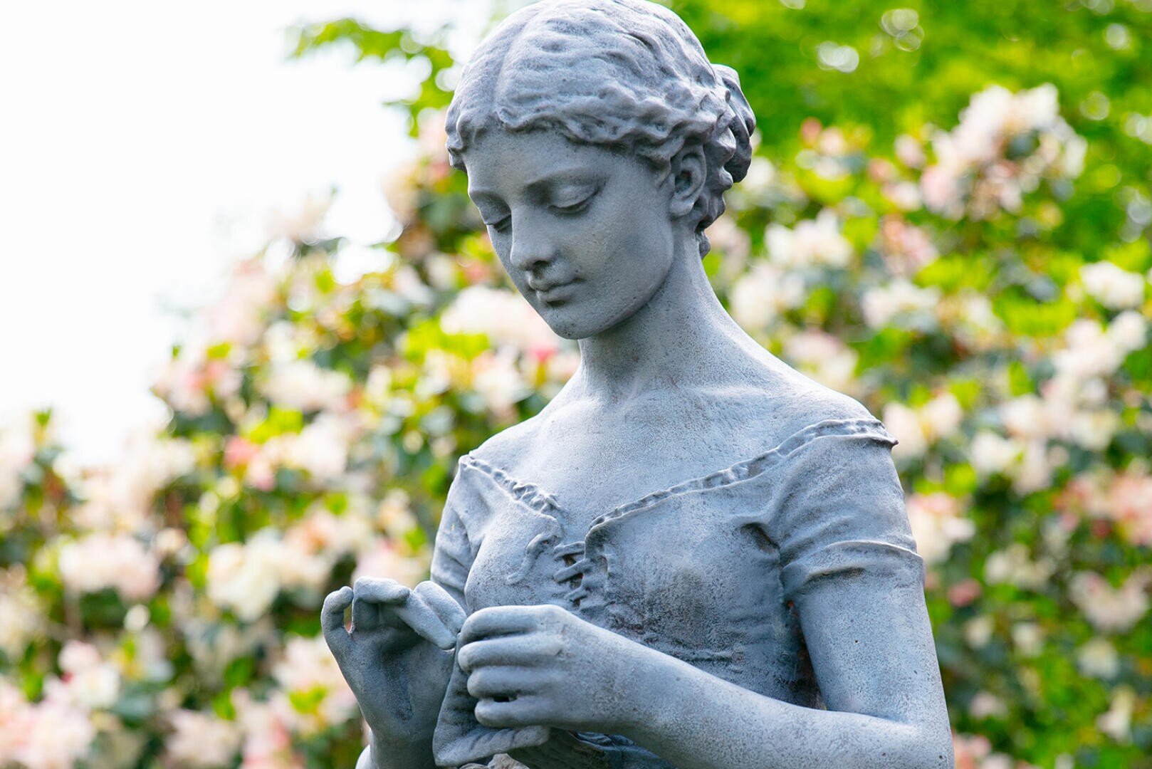 Nahaufnahme einer steinernen Statue einer jungen Frau mit geflochtenem Haar, die nach unten blickt und sanft einen kleinen Gegenstand hält, vor einem Hintergrund aus blühenden Büschen und grünem Laub.
