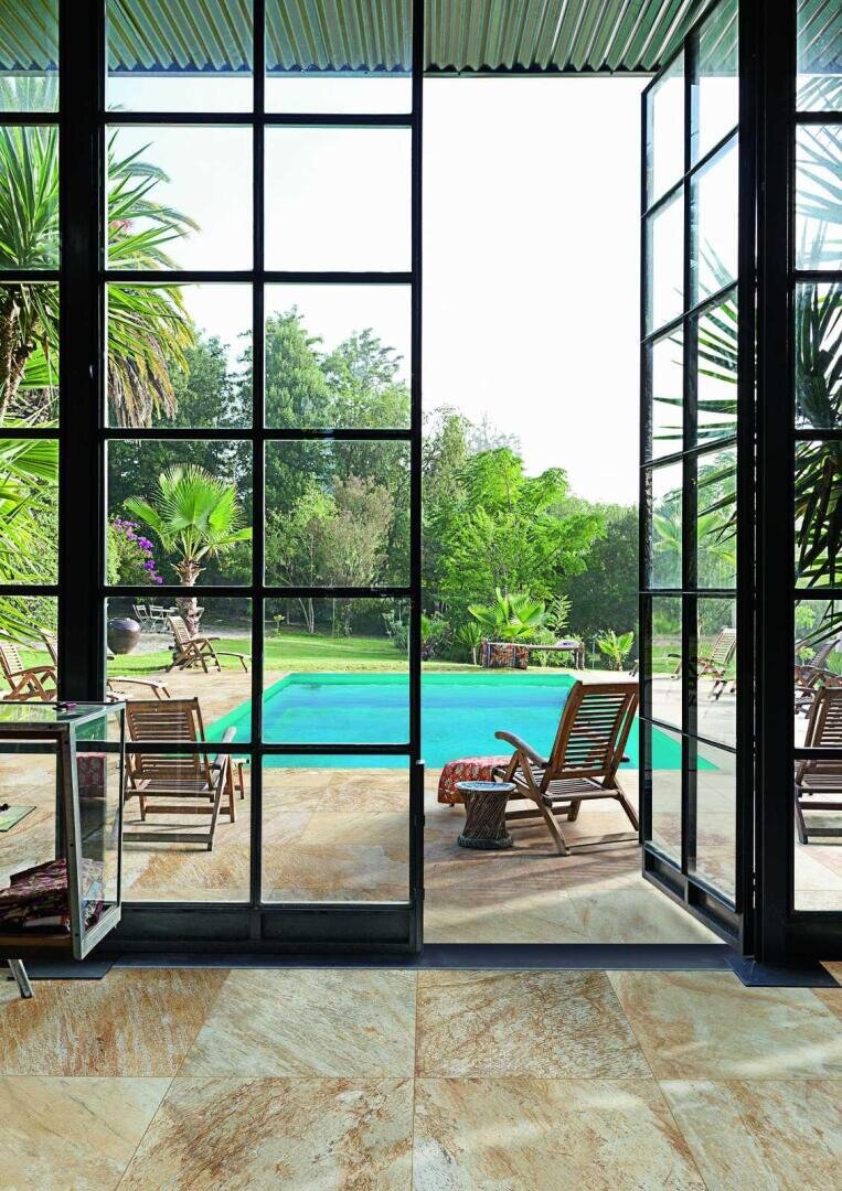 Large glass doors open to a patio with wooden lounge chairs and a table, overlooking a swimming pool surrounded by lush green trees and tropical plants under a bright sky.