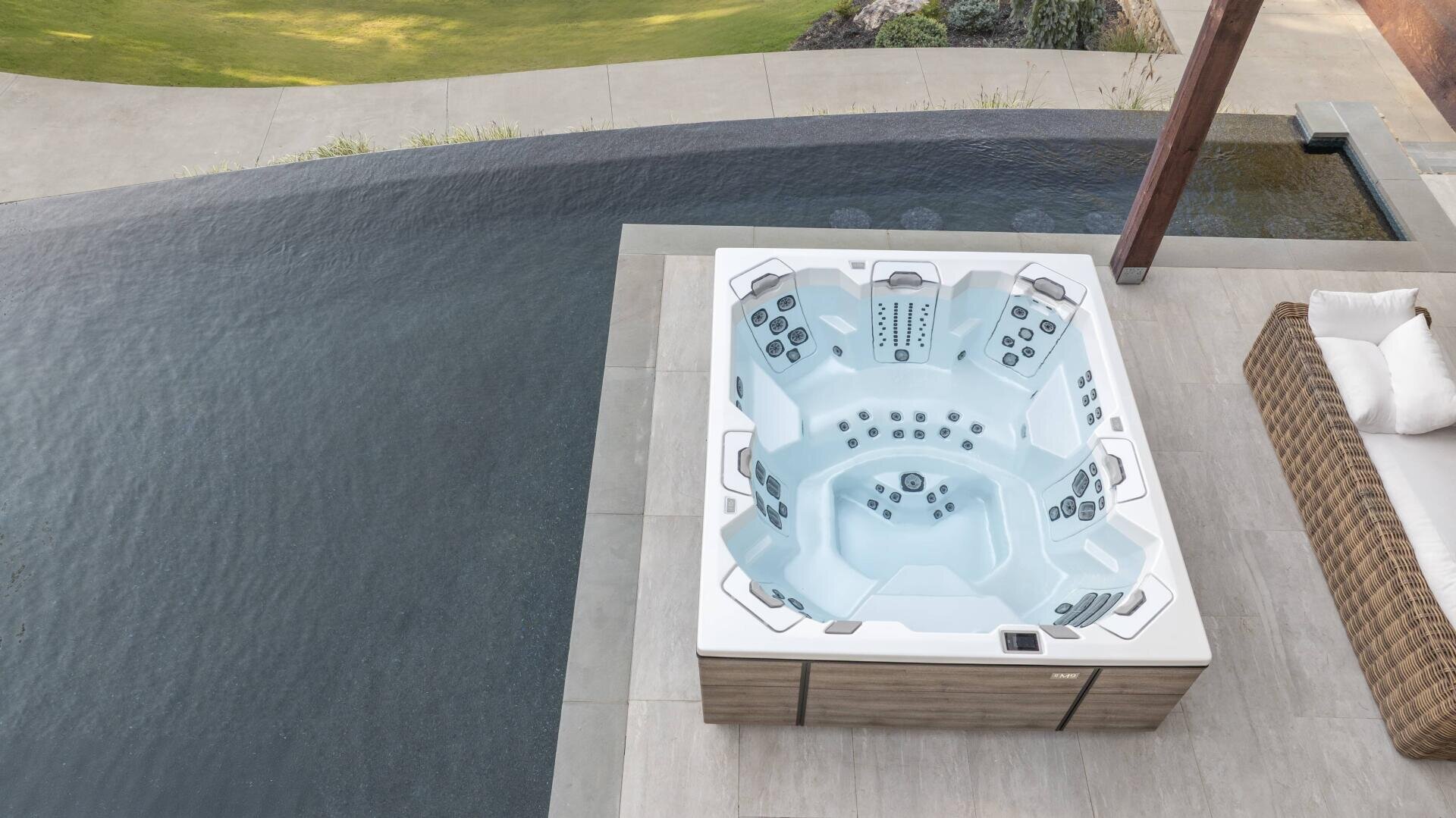 Ein moderner Whirlpool mit mehreren Sitzplätzen befindet sich neben einem Infinity-Pool auf einer gefliesten Terrasse, neben einem Korbsofa mit weißen Kissen und Blick auf das grüne Gras dahinter.