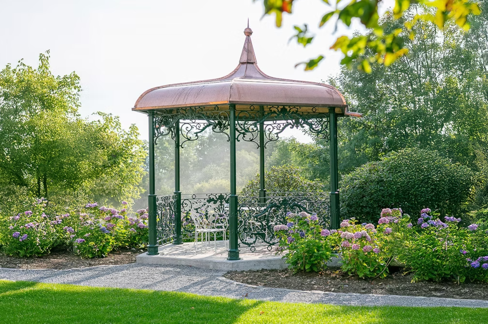 A decorative metal gazebo with a copper roof stands in a garden, surrounded by blooming purple flowers and green shrubs, with a white bench inside and trees in the background.