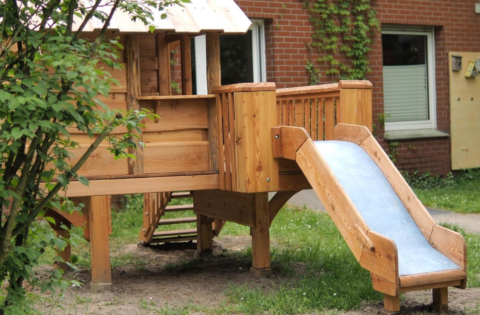 Ein hölzernes Spielhaus mit einer kleinen Veranda und einer blauen Rutsche steht in einem Hof mit Gras, Erde und einem grünen Busch in der Nähe, vor einem Backsteingebäude mit Fenstern.