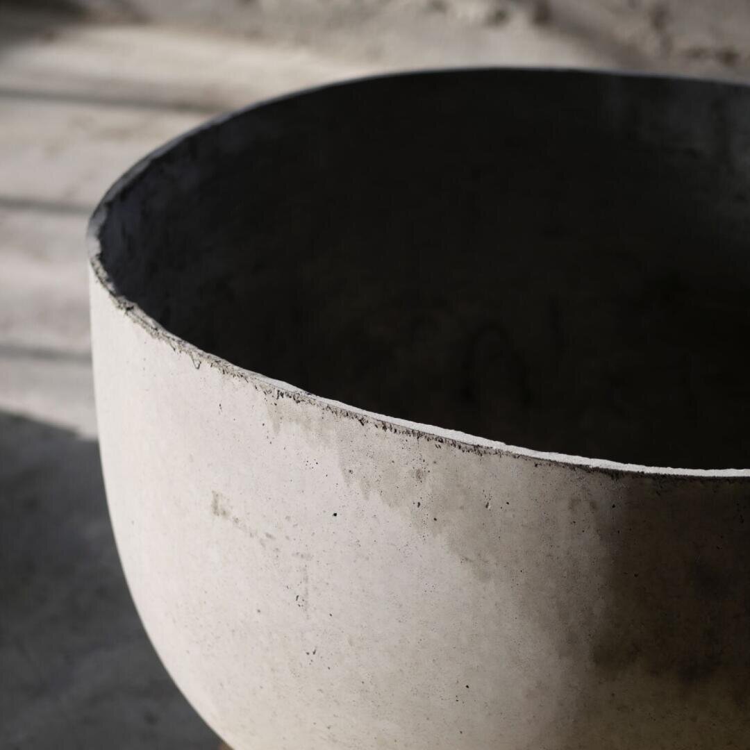 Nahaufnahme einer großen, runden, hellgrauen Betonschale mit rauem, unbearbeitetem Rand, die im natürlichen Licht auf einer glatten Oberfläche steht. Der Hintergrund ist unscharf.