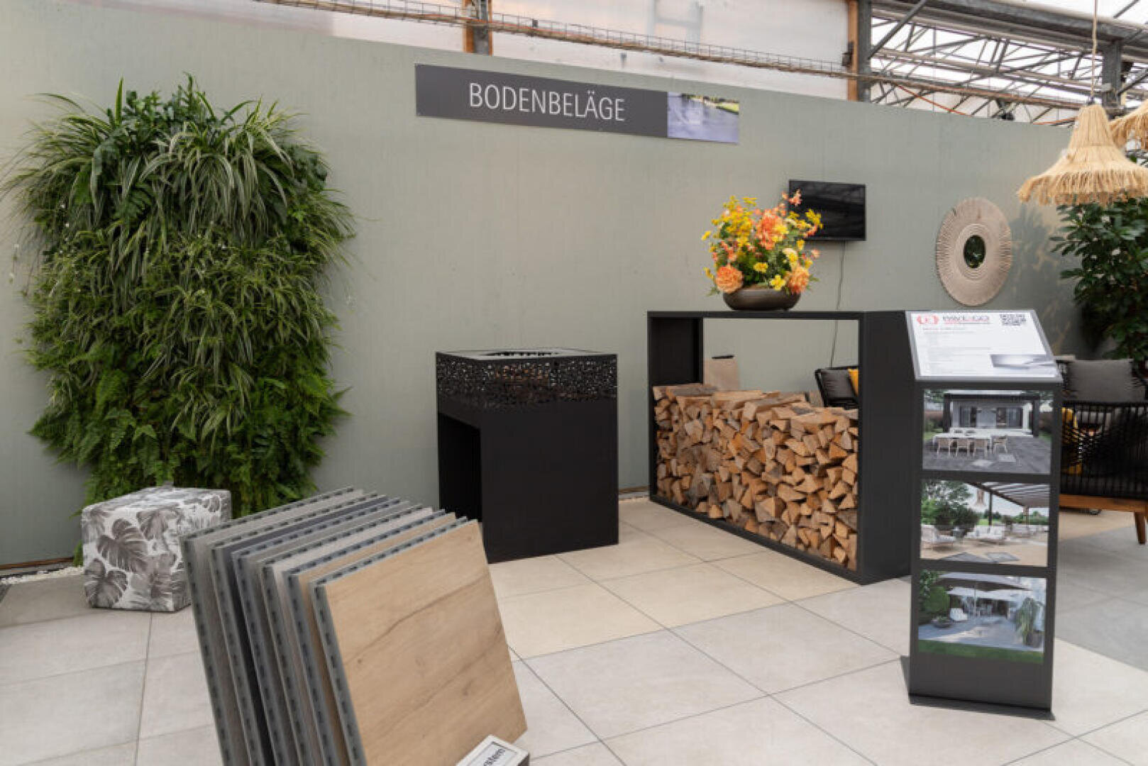 Ein Ausstellungsraum mit Mustern von Holzfußböden, einem Stapel Brennholz, einer dekorativen schwarzen Feuerstelle, einer Pflanzenwand, einer Blumenvase und einem Schild mit der Aufschrift BODENBELÄGE an einer hellgrauen Wand.