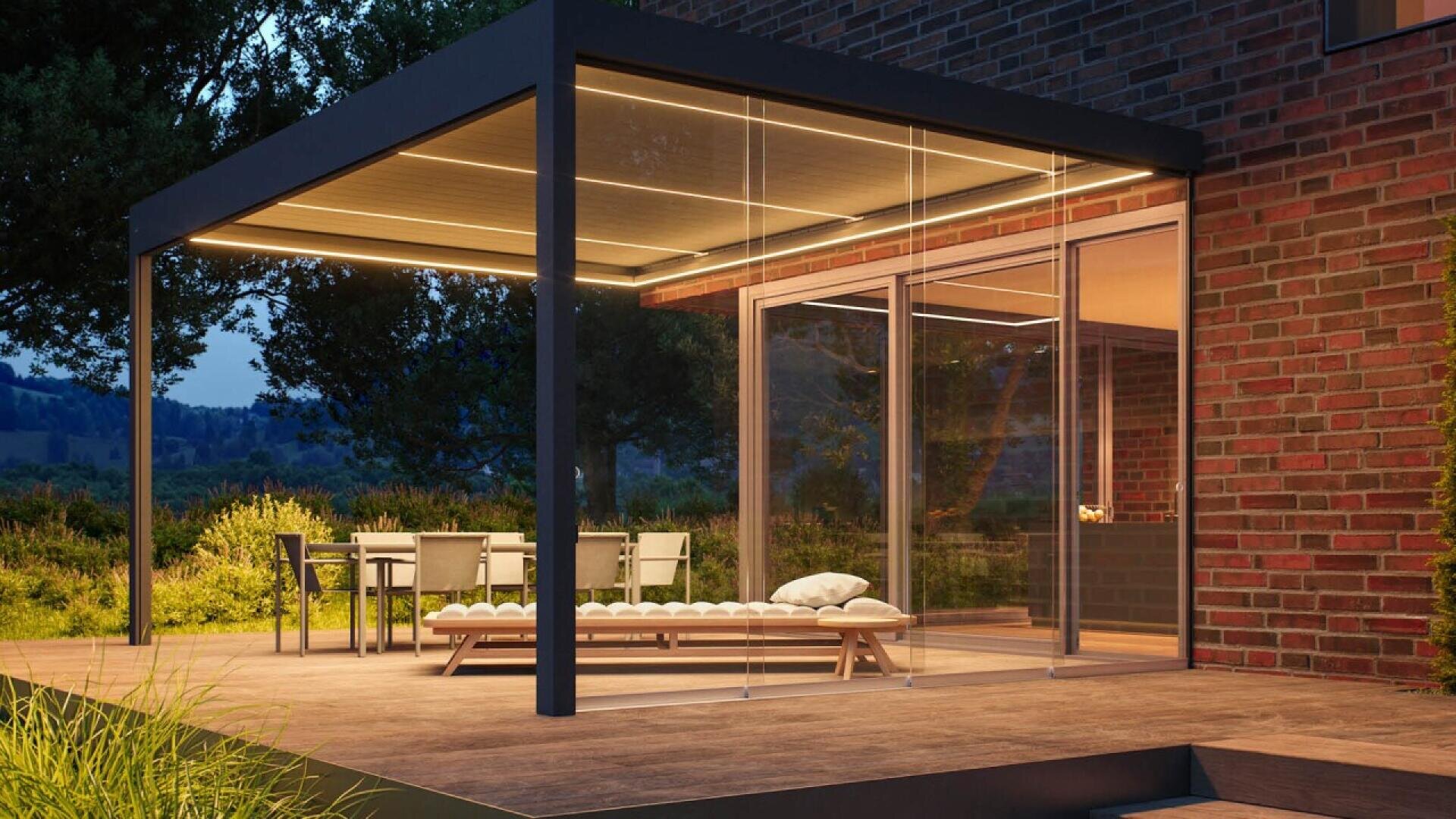 A modern patio with a sleek pergola, glass walls, and warm ceiling lights. There’s a dining table with chairs and a cushioned lounge chair on a wooden deck next to a brick house, surrounded by greenery at dusk.