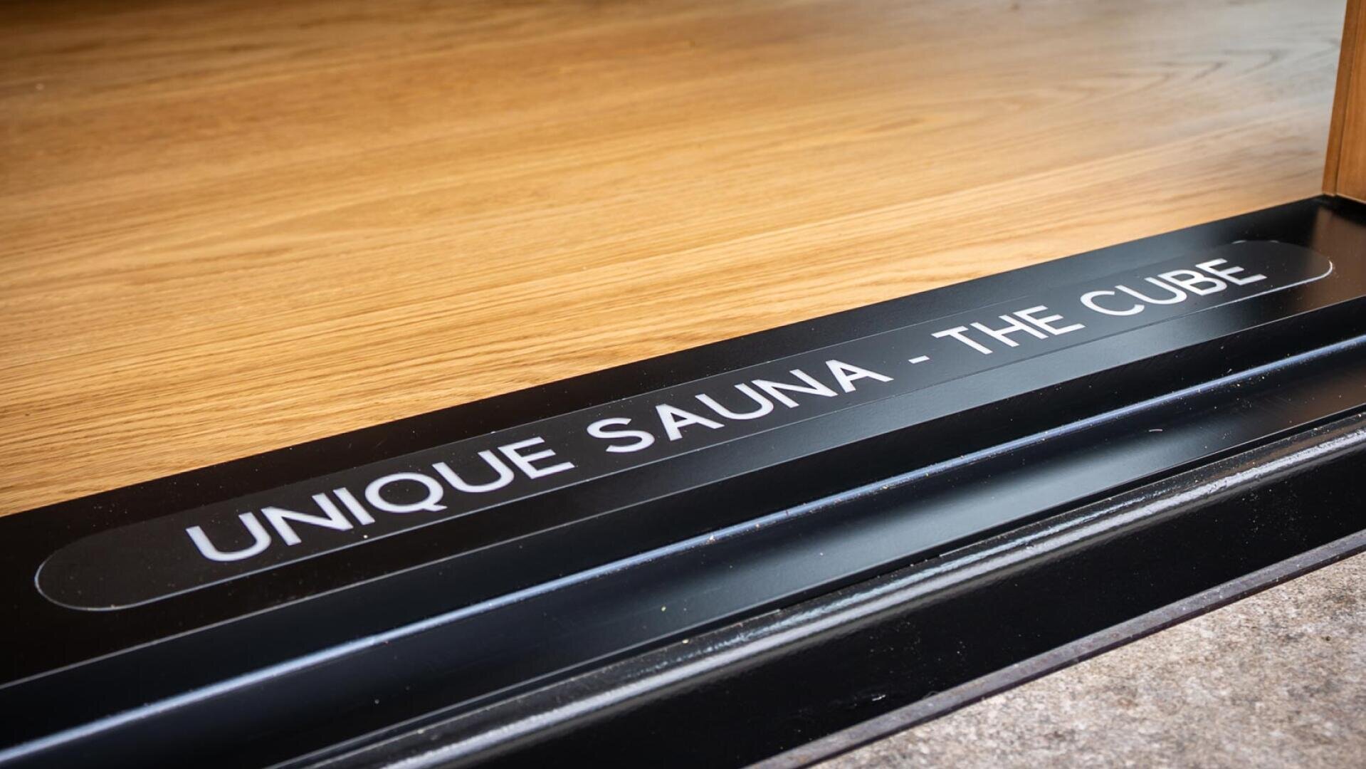 Schwarze Türschwelle mit weißer Aufschrift UNIQUE SAUNA - THE CUBE am Eingang zu einem Raum mit hellem Holzfußboden.
